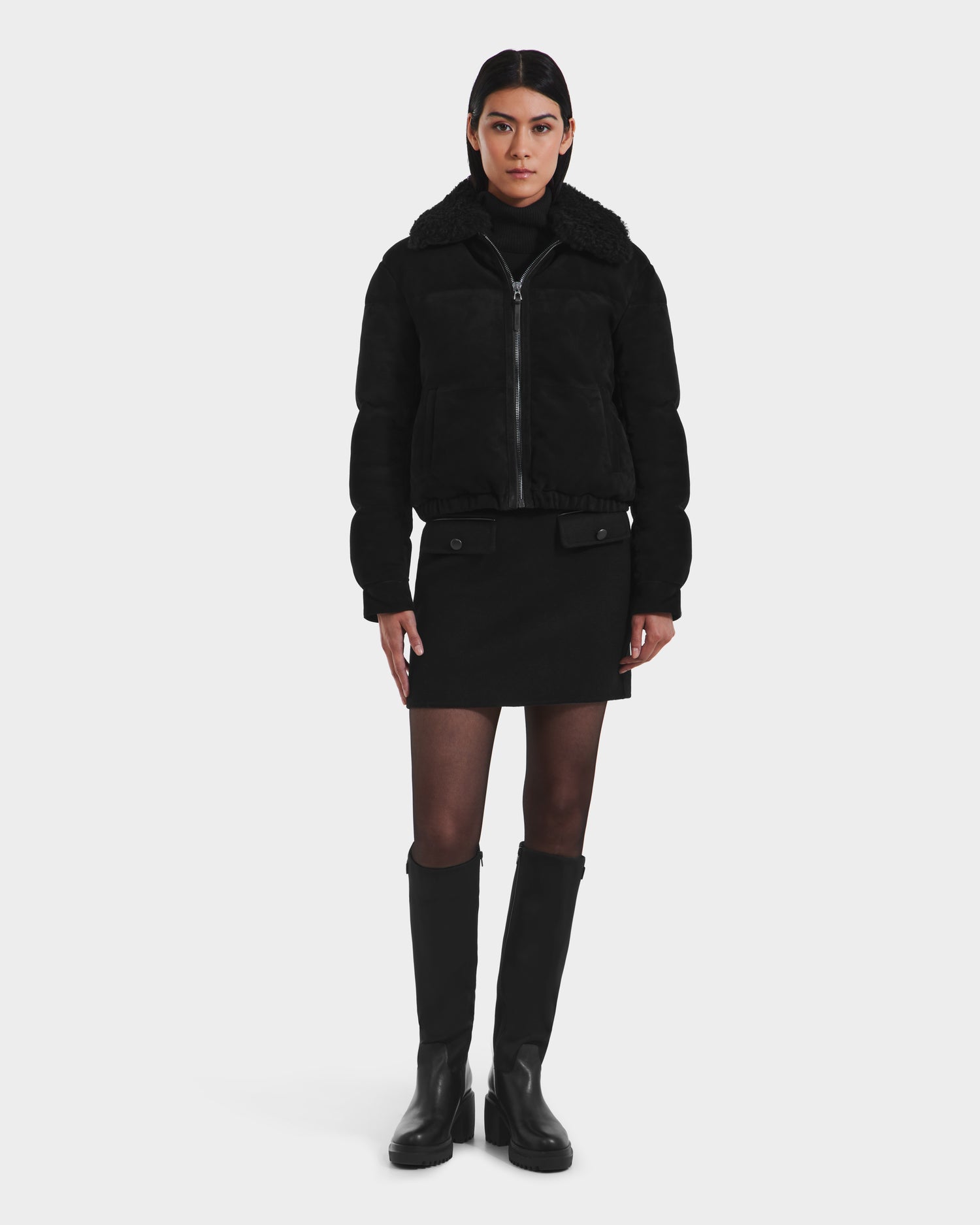 A woman with straight dark hair and a neutral expression faces forward against a white background, wearing the Rudsak STACEY - 8125049 Black womens aviator jacket with removable shearling collar, black mini skirt, sheer tights, and knee-high black boots.