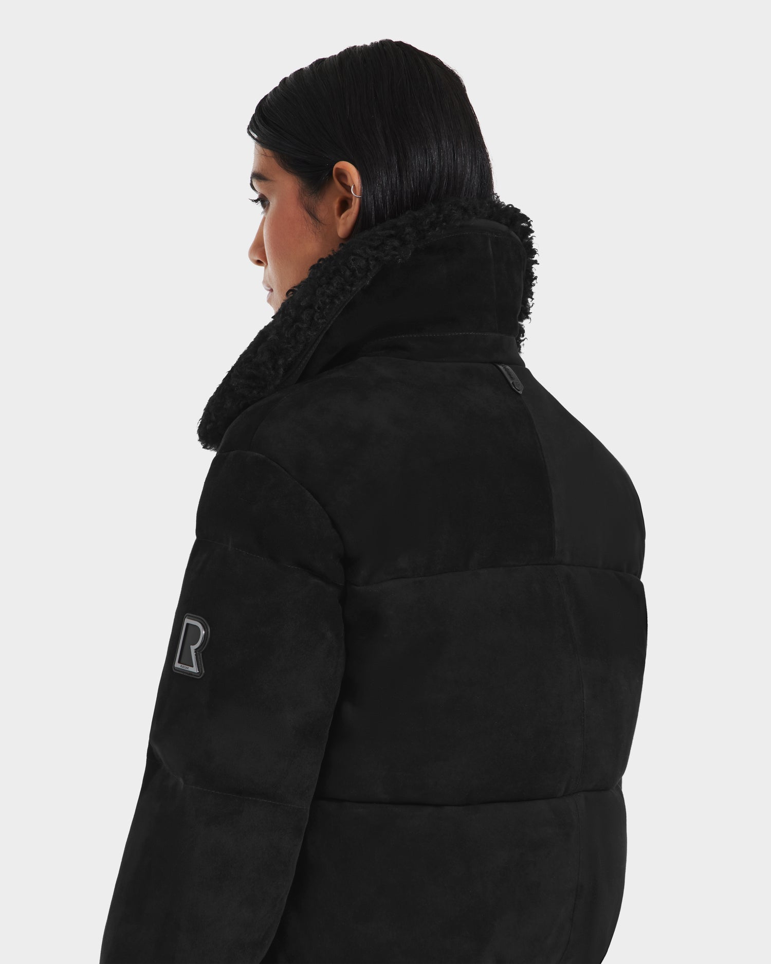 A person with dark hair wears the Rudsak STACEY - 8125049 Black women’s aviator jacket, featuring timeless style and a high sherpa-lined collar—seen from the back and side against a plain light background.
