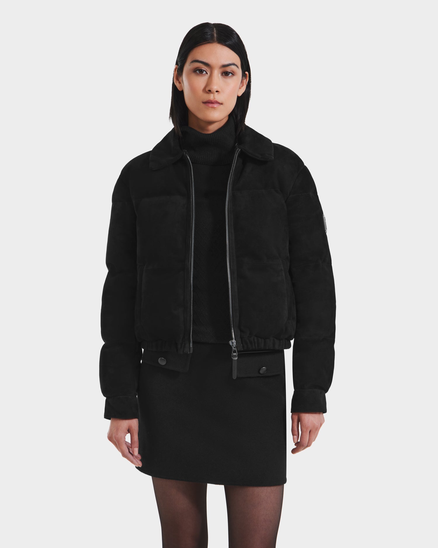 A woman with straight dark hair wears the Rudsak STACEY - 8125049 Black aviator jacket with a removable shearling collar over a black turtleneck sweater and skirt, standing against a plain white background.