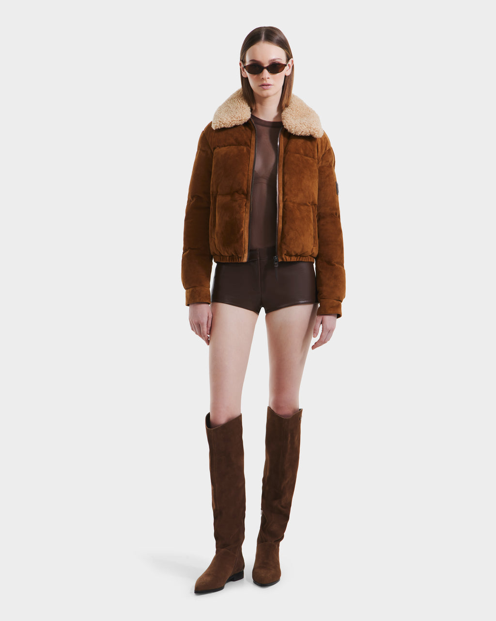 A woman wearing the Rudsak STACEY - 8125049 Mocha aviator jacket with a shearling collar, brown top, dark shorts, knee-high boots, and sunglasses stands against a plain white background.