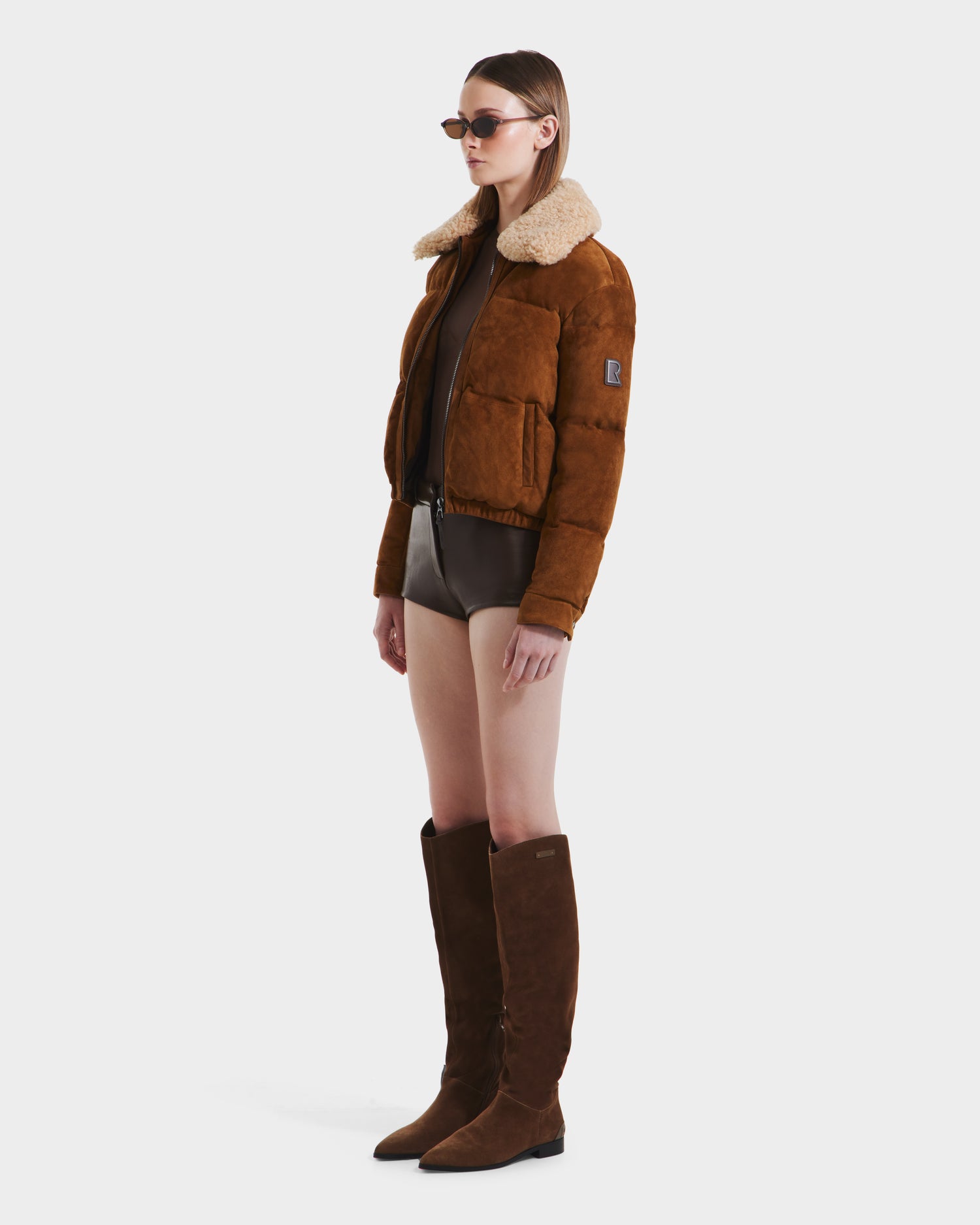 A woman in Rudsak’s STACEY - 8125049 Mocha womens aviator jacket with a shearling collar, brown bodysuit, sunglasses, and knee-high brown suede boots stands against a plain white background.