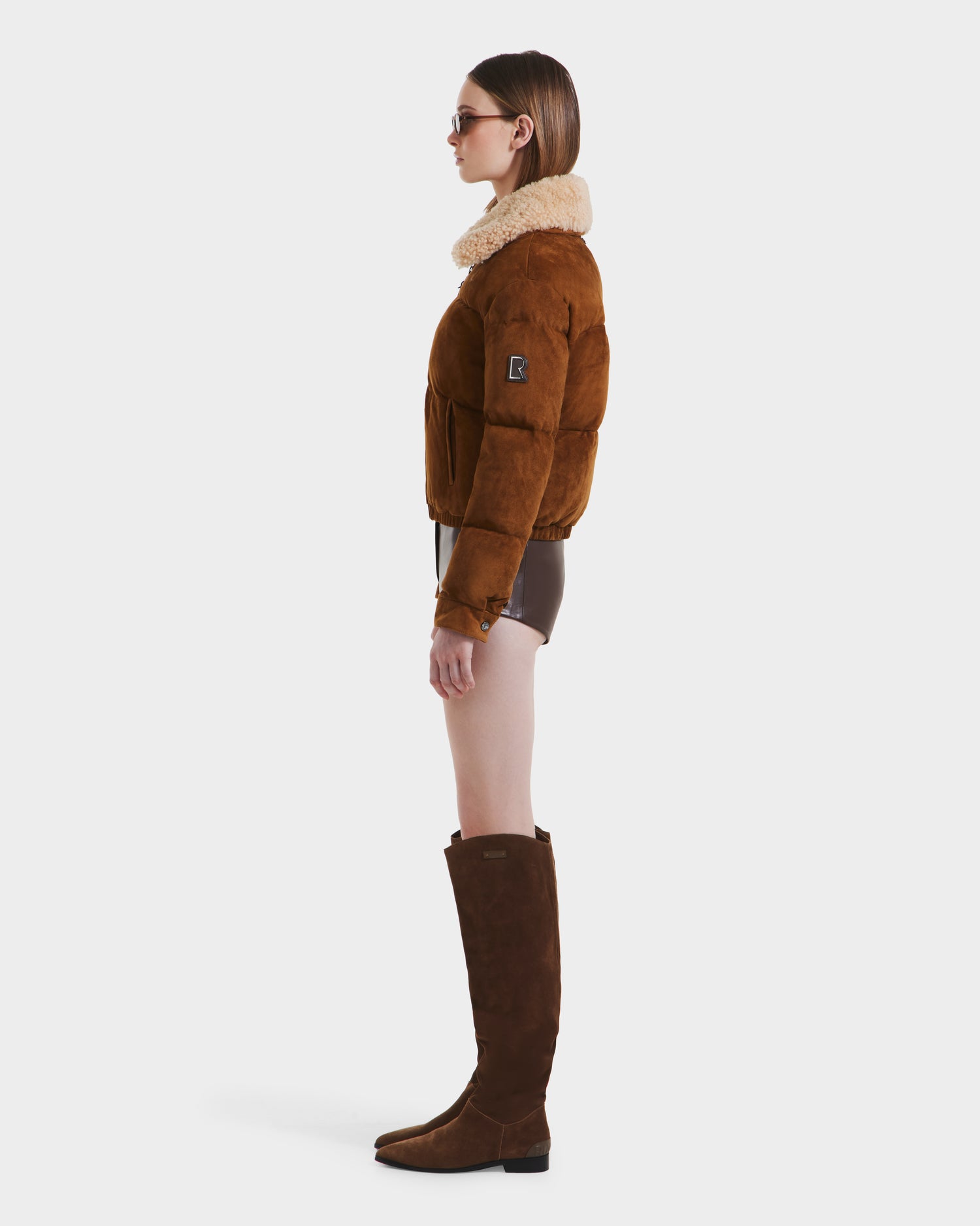 A woman poses in profile against a white background, wearing Rudsaks STACEY - 8125049 Mocha suede bomber jacket with a shearling collar, brown shorts, tall suede boots, sunglasses, and straight hair.
