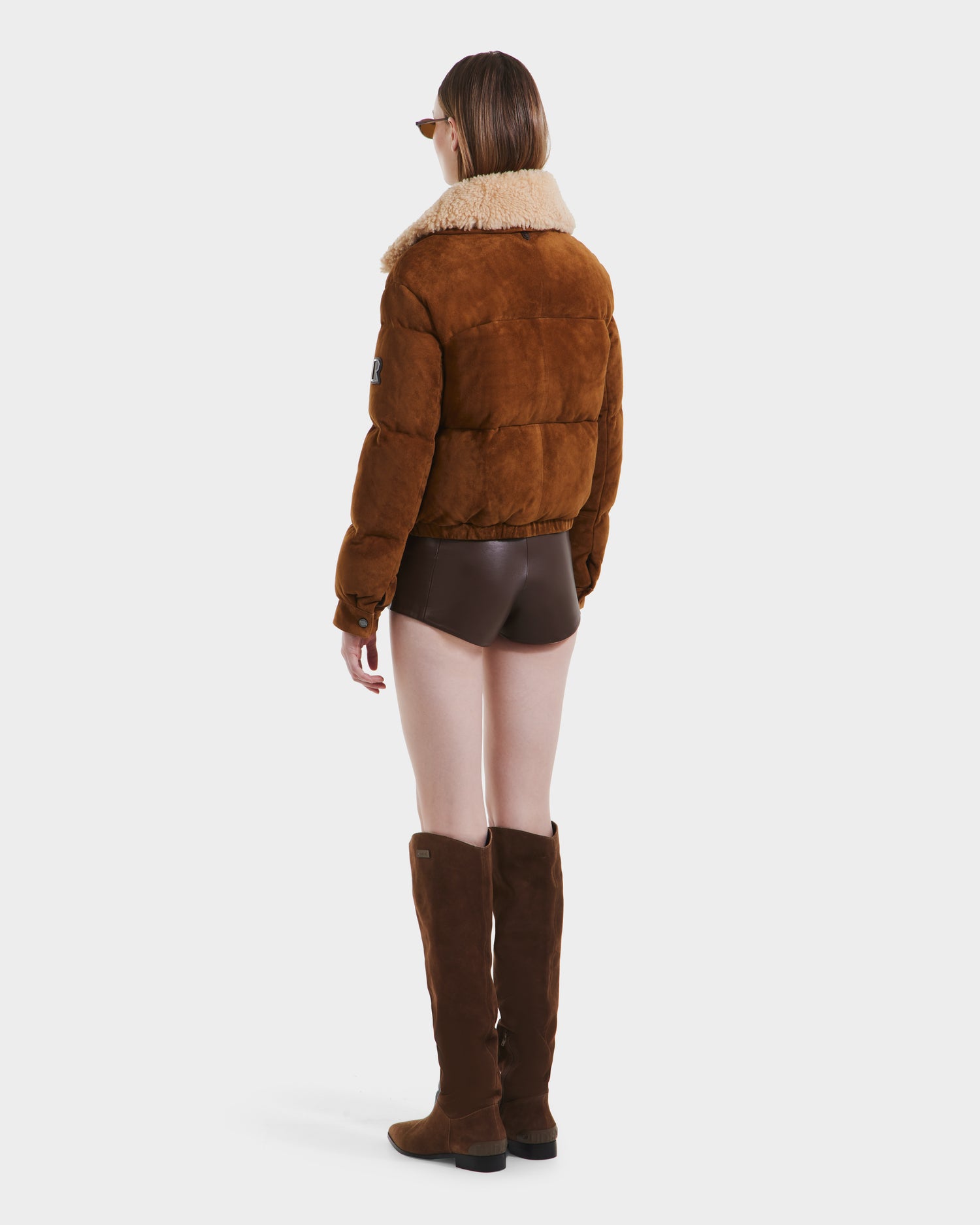 A person stands facing away in the Rudsak STACEY - 8125049 Mocha suede bomber jacket with shearling collar, paired with brown high-waisted shorts, knee-high boots, and sunglasses against a plain white background.