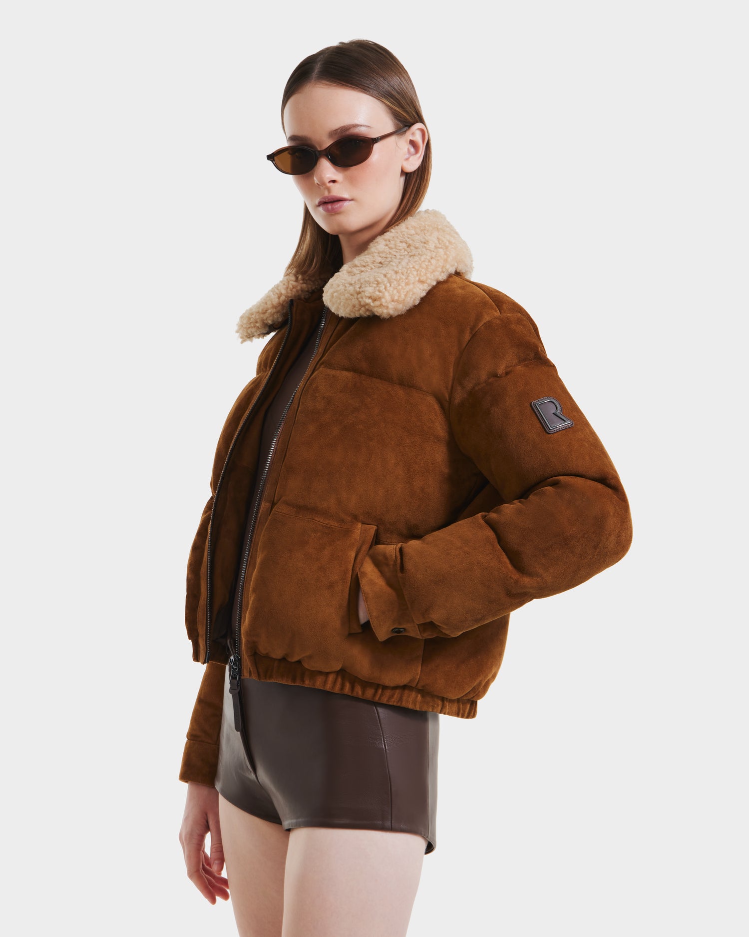 A woman wears Rudsak's STACEY - 8125049 Mocha aviator jacket with a shearling collar, brown sunglasses, and a brown bodysuit while posing against a plain white background.