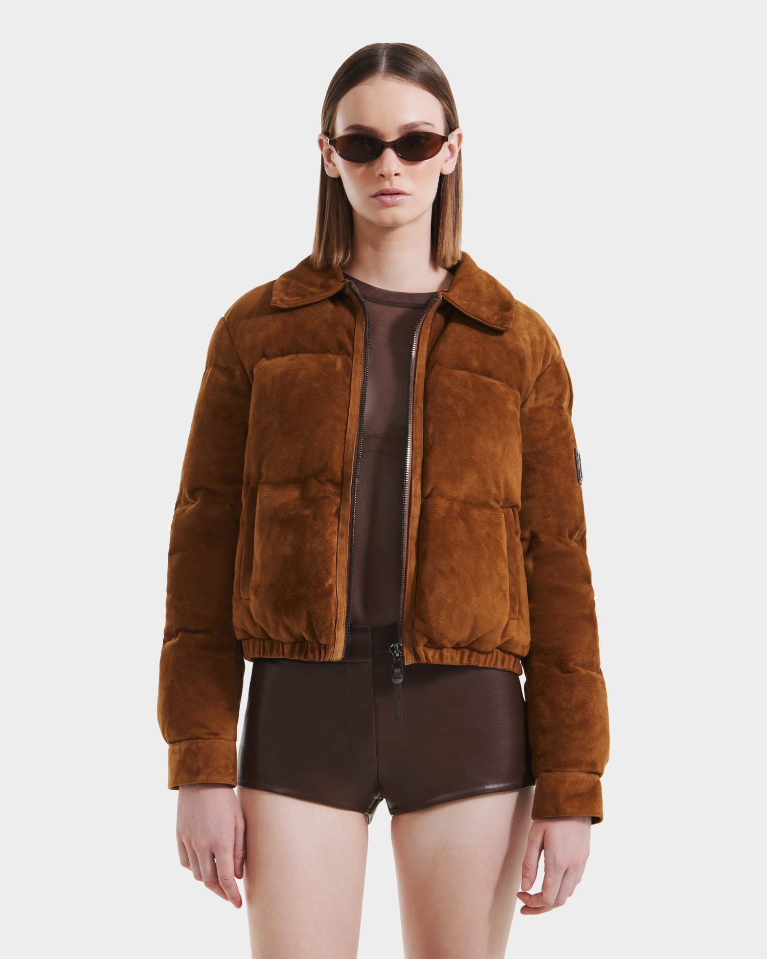 A woman with straight brown hair wears Rudsaks STACEY - 8125049 Mocha sunglasses, a suede bomber jacket with a shearling collar, sheer brown top, and high-waisted shorts against a plain white background.