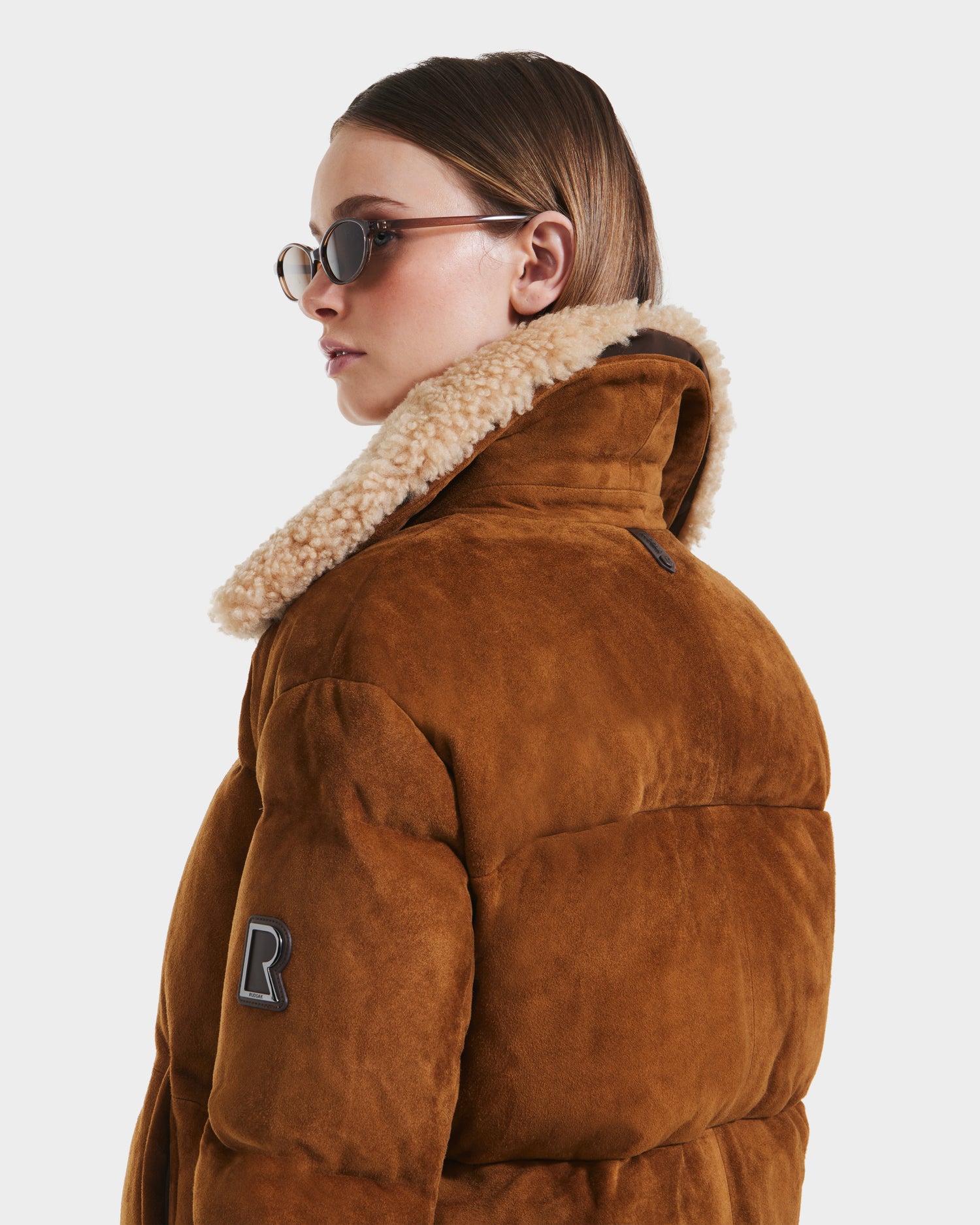 A woman in the Rudsak STACEY - 8125049 Mocha women’s aviator jacket with a shearling collar and an “R” patch on the sleeve, plus oval sunglasses, looks to the side against a plain background.
