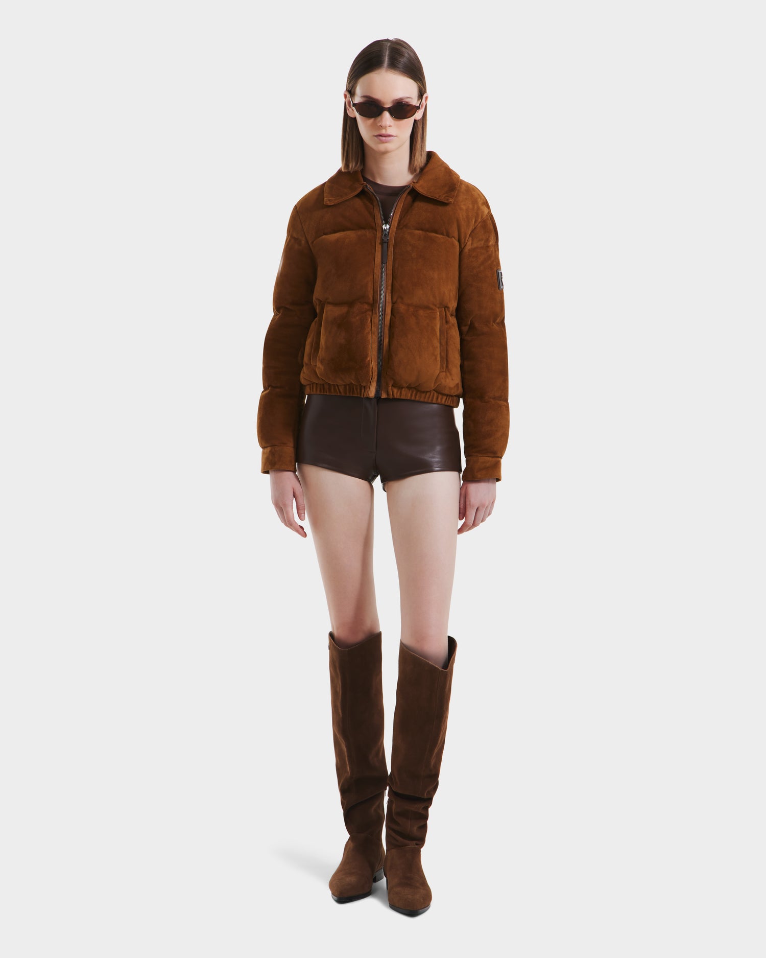 A person wears Rudsak’s STACEY - 8125049 Mocha women’s aviator jacket with a shearling collar, sunglasses, dark brown shorts, and tall brown suede boots, standing against a plain white background.