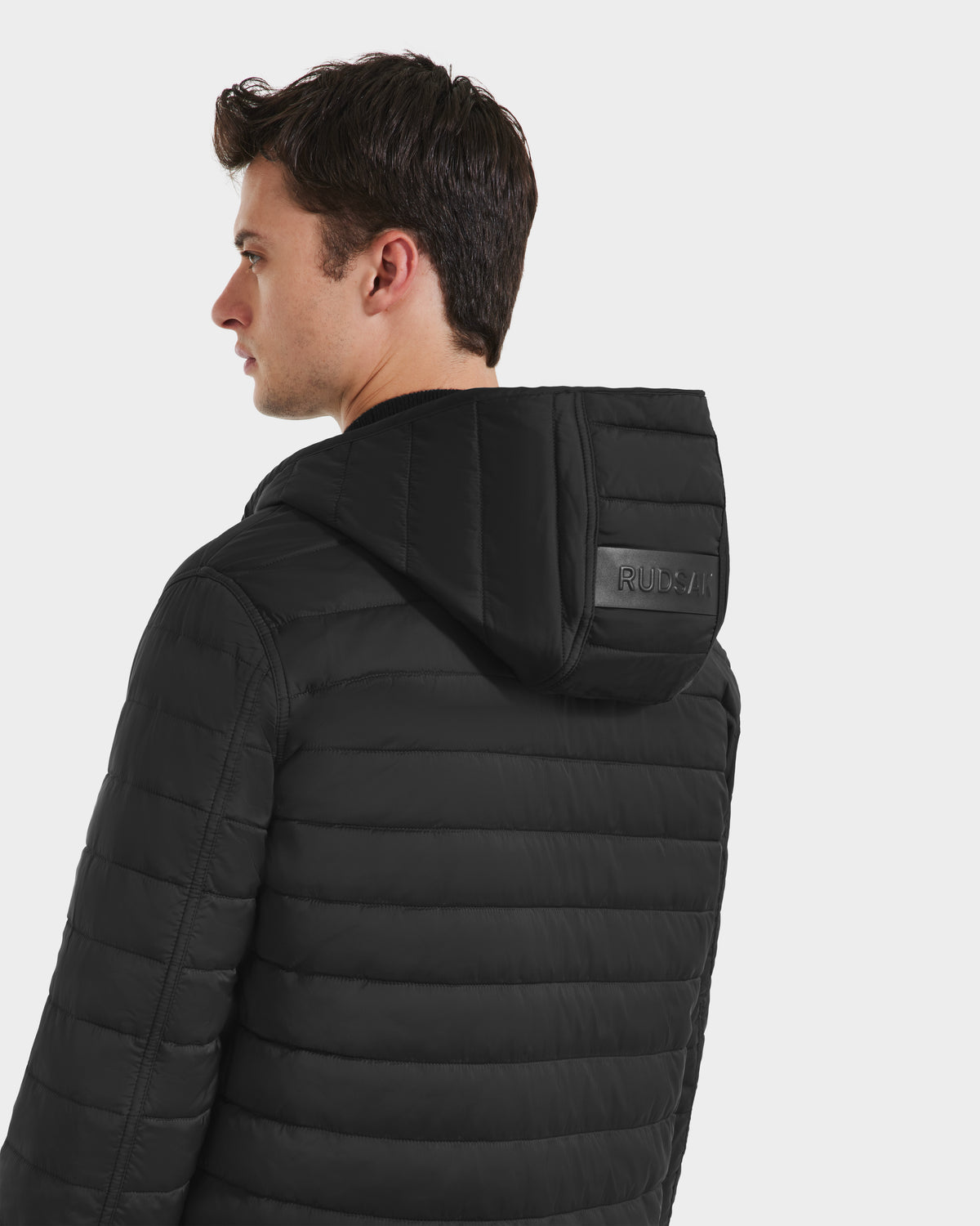 A young man with short brown hair models the Rudsak STANTON - 6125531 Black jacket, turning left to display its weather-resistant, quilted design and the Rudsak logo embossed on the hood.