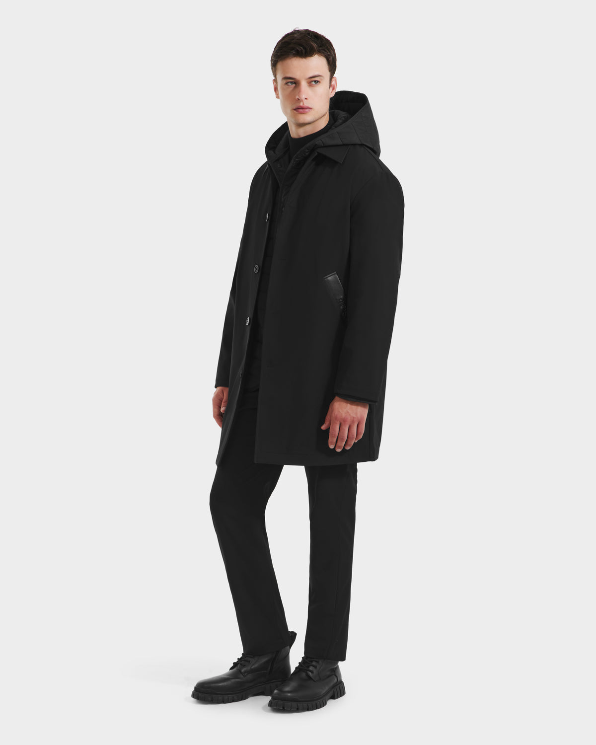 A man stands slightly turned against a plain white background, wearing the Rudsak STANTON - 6125531 Black weather-resistant hooded coat, paired with black pants and black shoes.