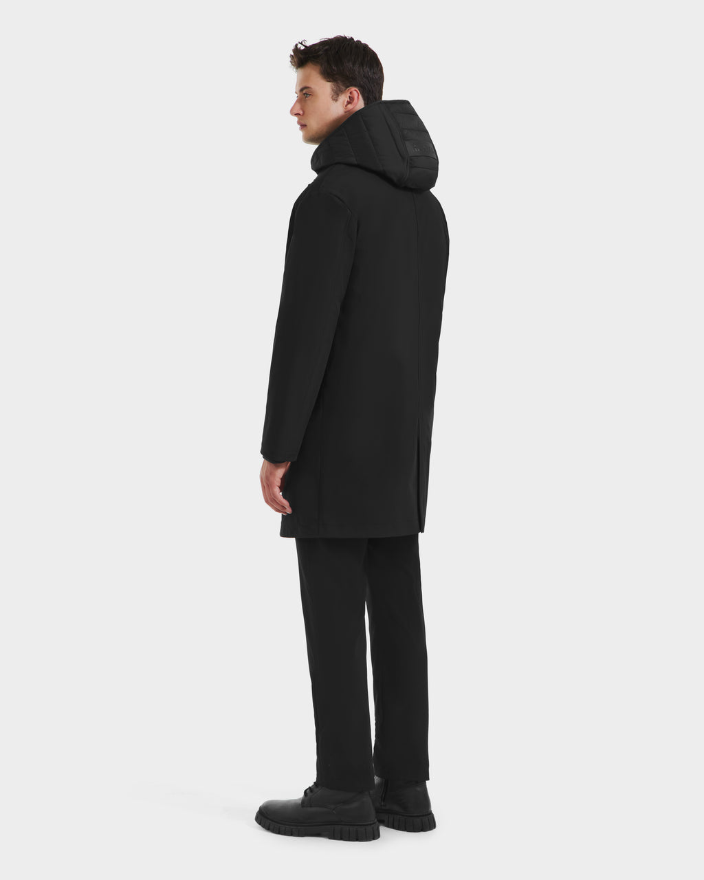 A man wearing the Rudsak STANTON - 6125531 Black overcoat, paired with black pants and shoes, stands facing away with his head turned slightly to the side against a plain white background.
