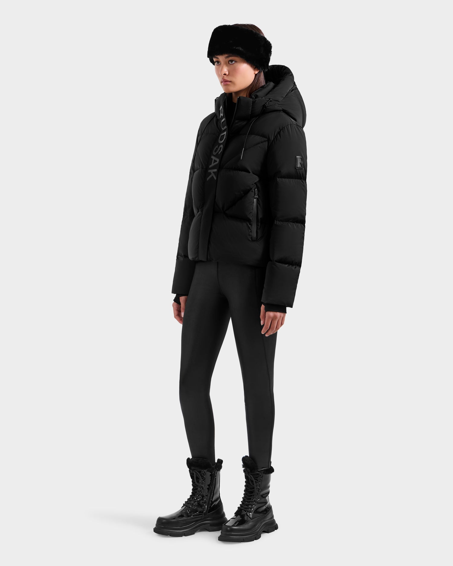 A person wearing the Rudsak STEFANIE M - L125526 Black puffer jacket with a hood, black leggings, winter boots, and a fur hat stands against a plain white background.