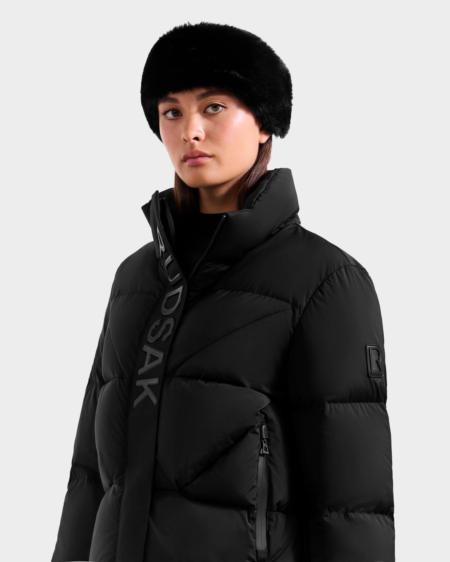 A woman wears the Rudsak STEFANIE M - L125526 Black puffer jacket and a black fur hat, standing against a plain light gray background.