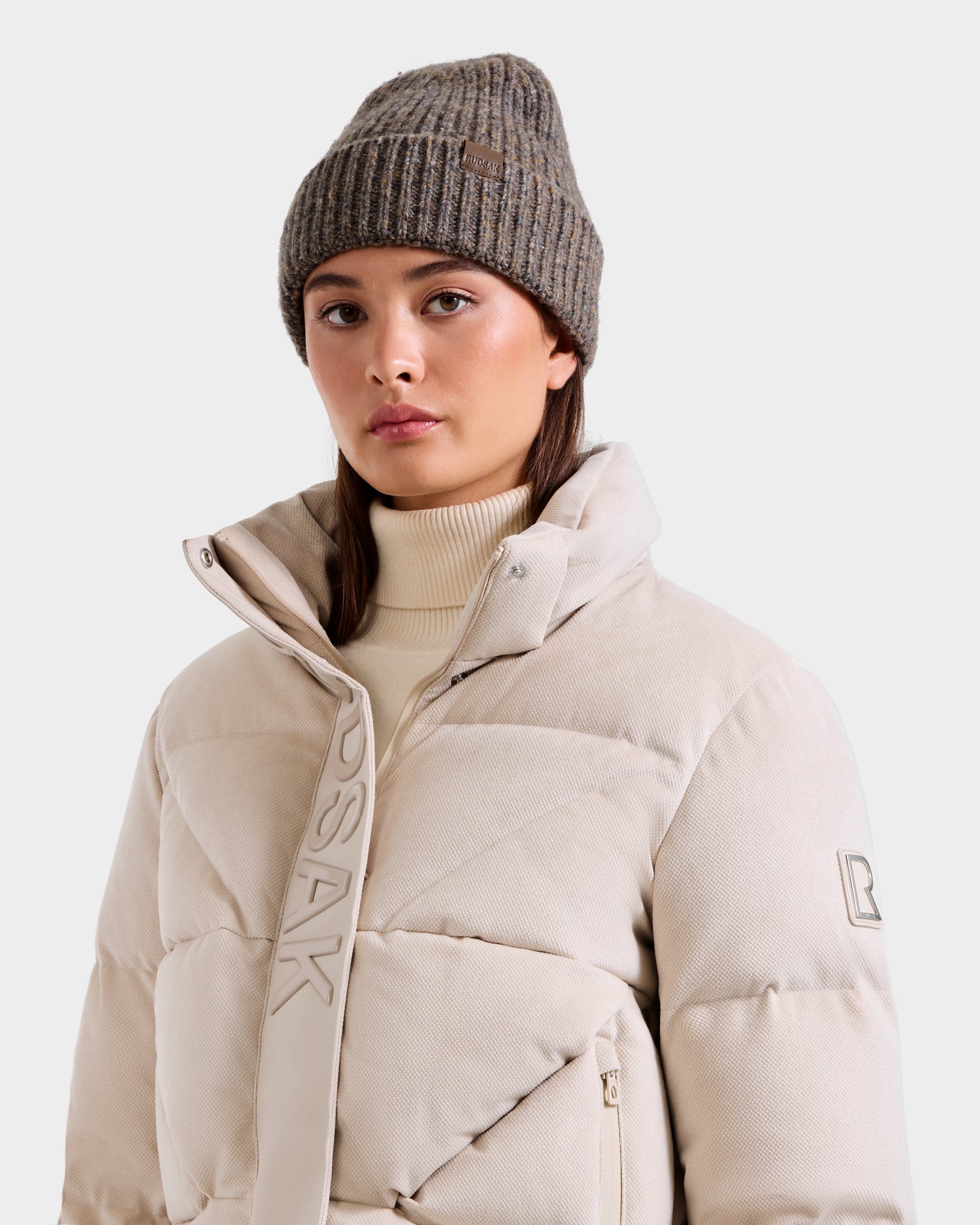 A woman wears the Rudsak STEFANIE V - L125584 Vanilla down puffer jacket with a high collar and light beige turtleneck, paired with a ribbed gray knit beanie—ideal for any winter wardrobe.