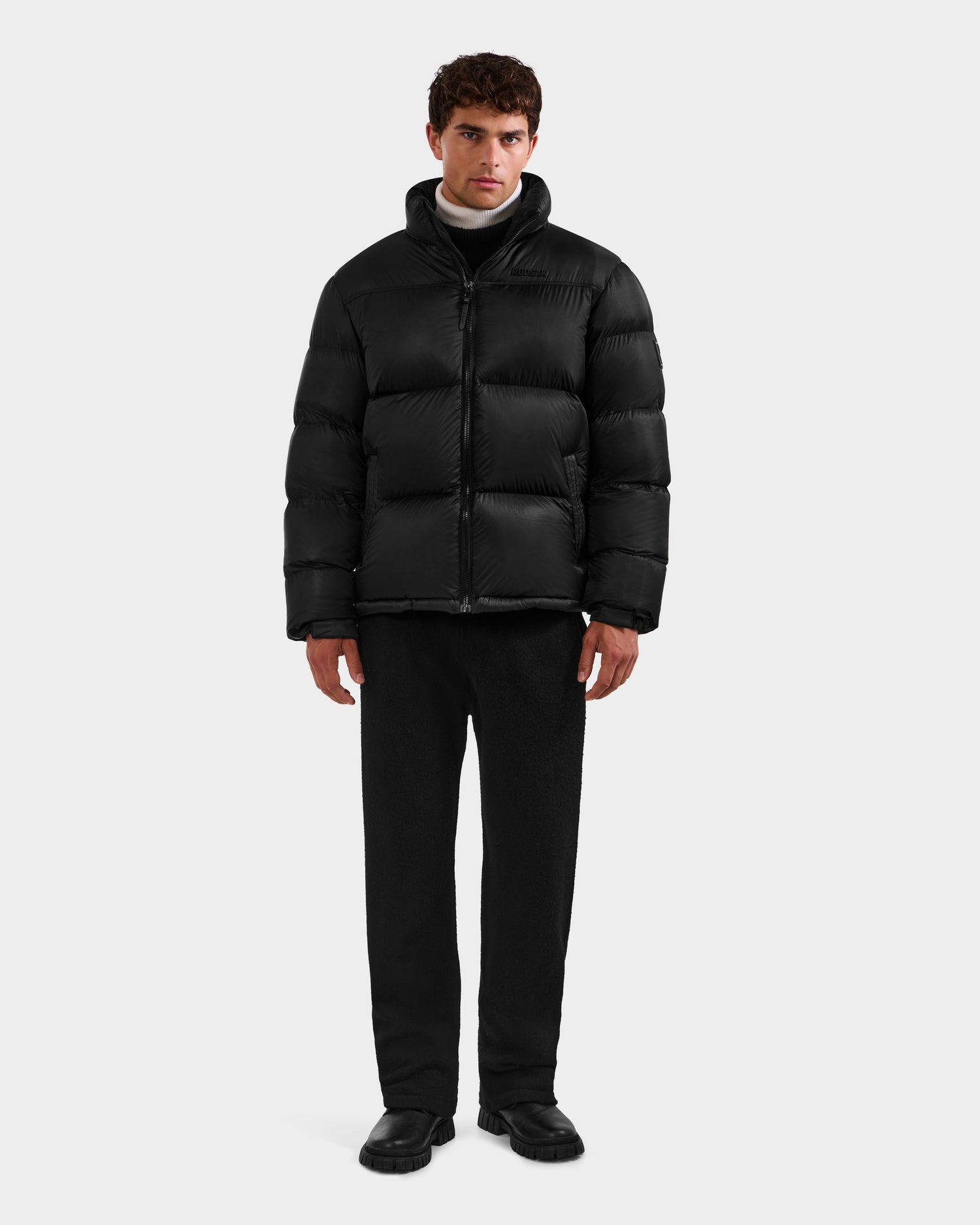 A man stands facing forward against a plain white background, wearing the Rudsak STEFANO - 6125574 Black men’s down puffer winter jacket, paired with black pants and black shoes.