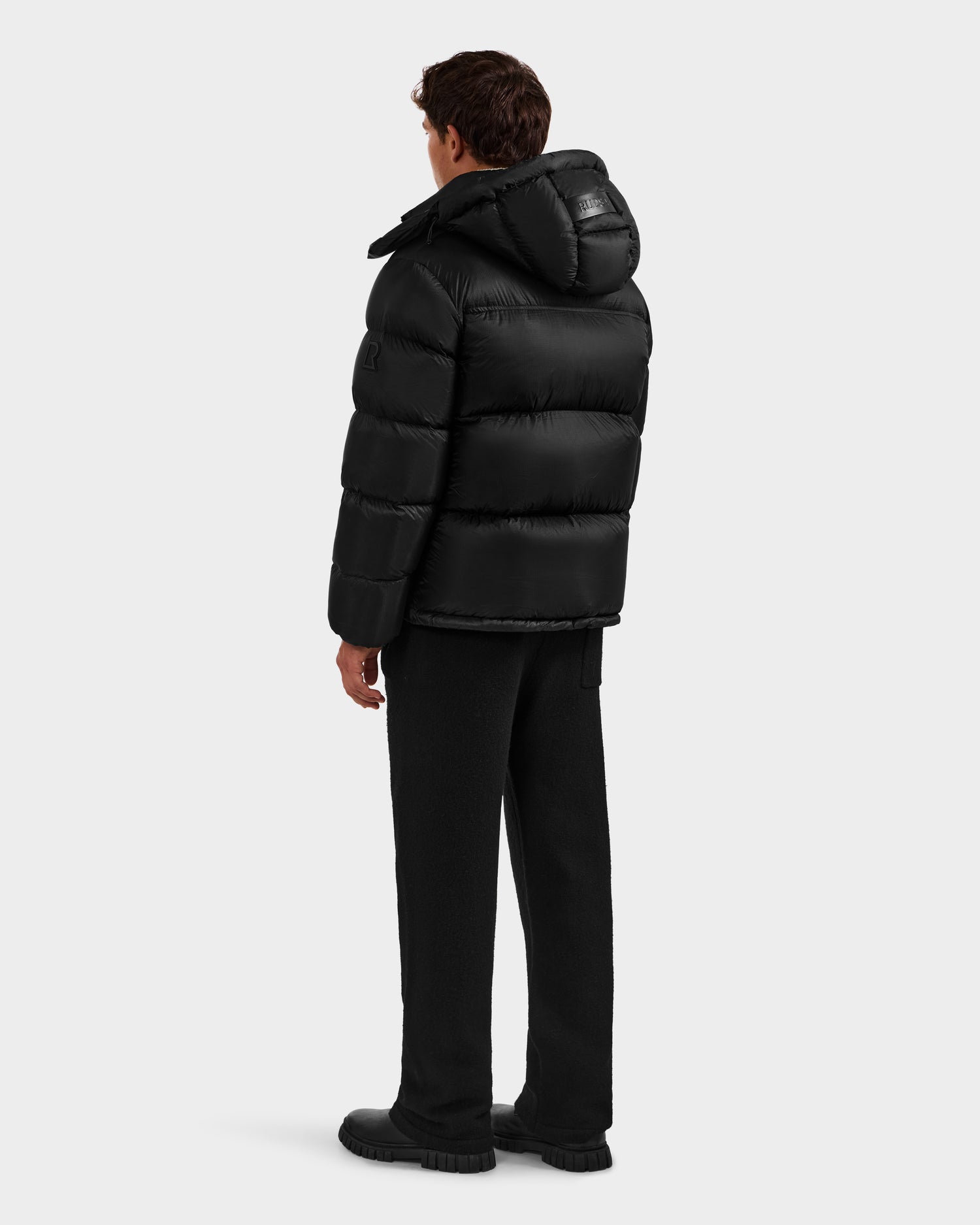 A person faces away, wearing the Rudsak STEFANO - 6125574 Black men’s down puffer jacket with a hood, black pants, and black shoes, set against a plain light gray background.