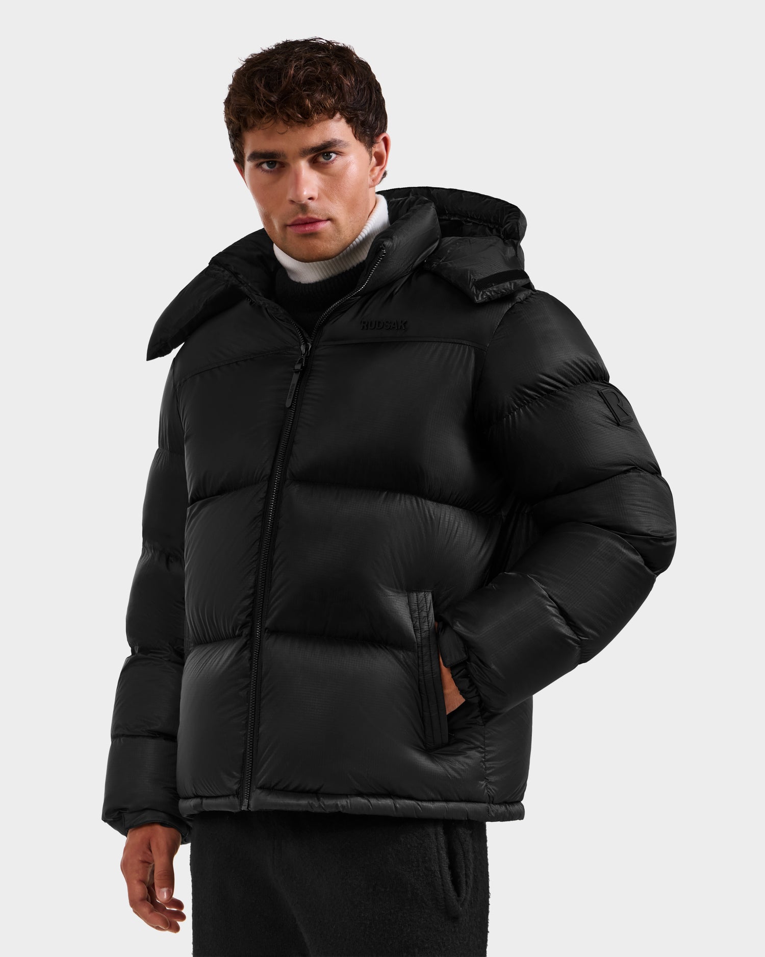 A man with short curly brown hair wears the Rudsak STEFANO - 6125574 Black men's down puffer jacket with a hood, RDS-certified down fill, standing against a light gray background and looking directly at the camera with a neutral expression.