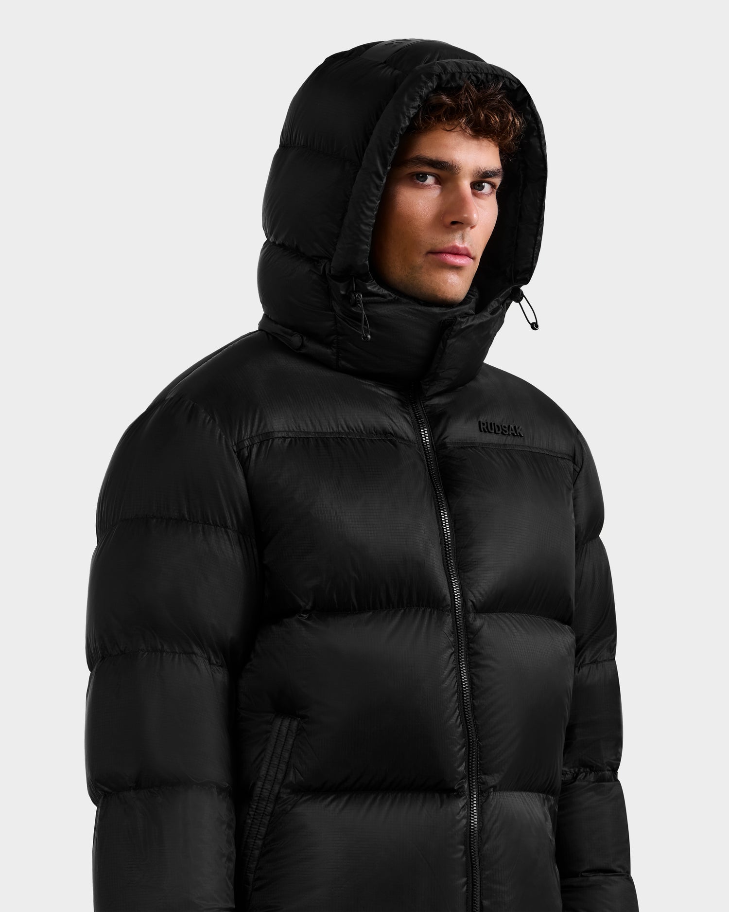A young man wearing the Rudsak STEFANO - 6125574 Black men’s down puffer with a high collar and horizontal quilting looks slightly to the side against a plain light gray background. This winter jacket features RDS-certified down for warmth.