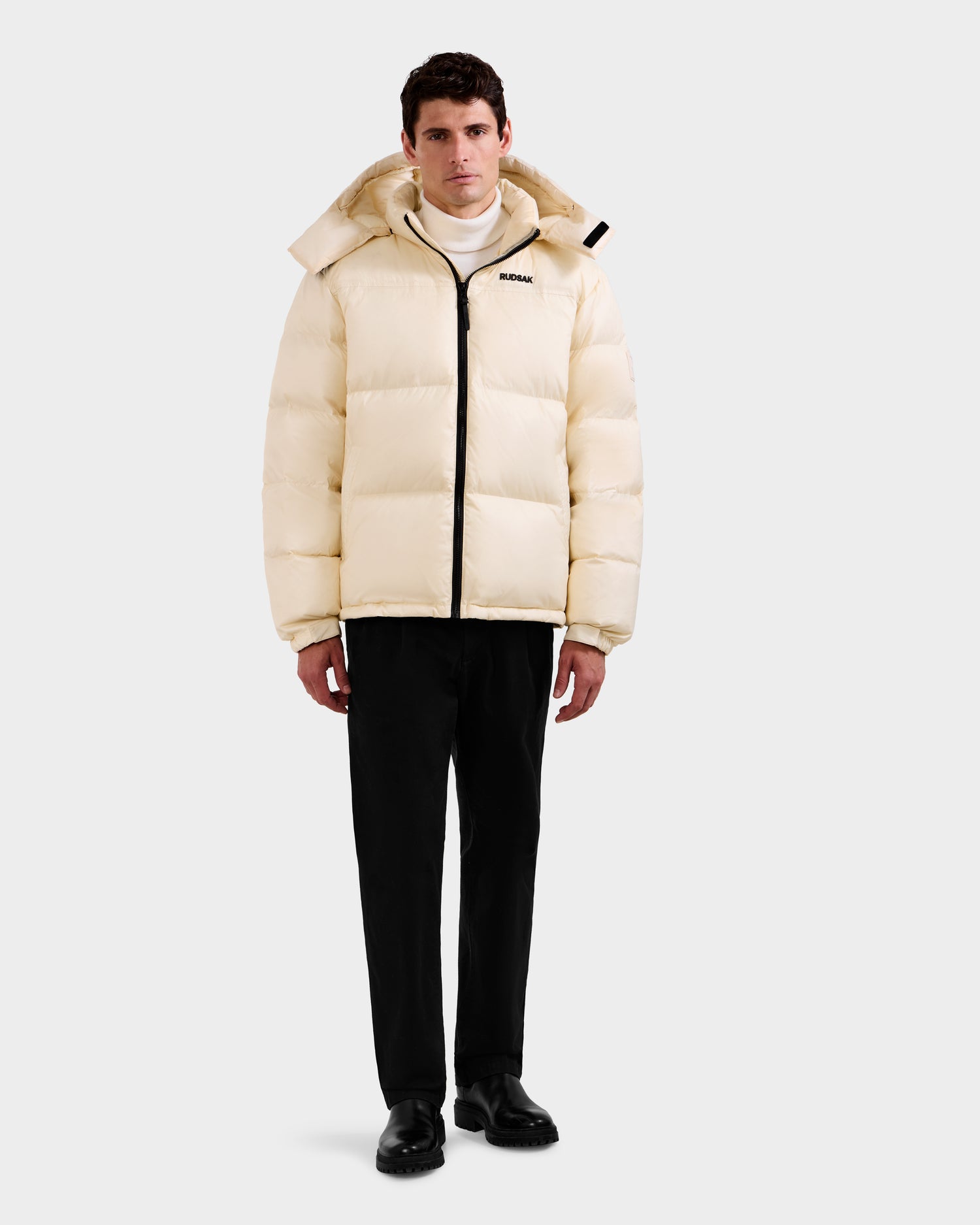 A man faces forward wearing the Rudsak STEFANO - 6125574 Vanilla quilted down jacket with a hood, black pants, black shoes, and a white turtleneck—an effortlessly stylish winter look against a plain light background.