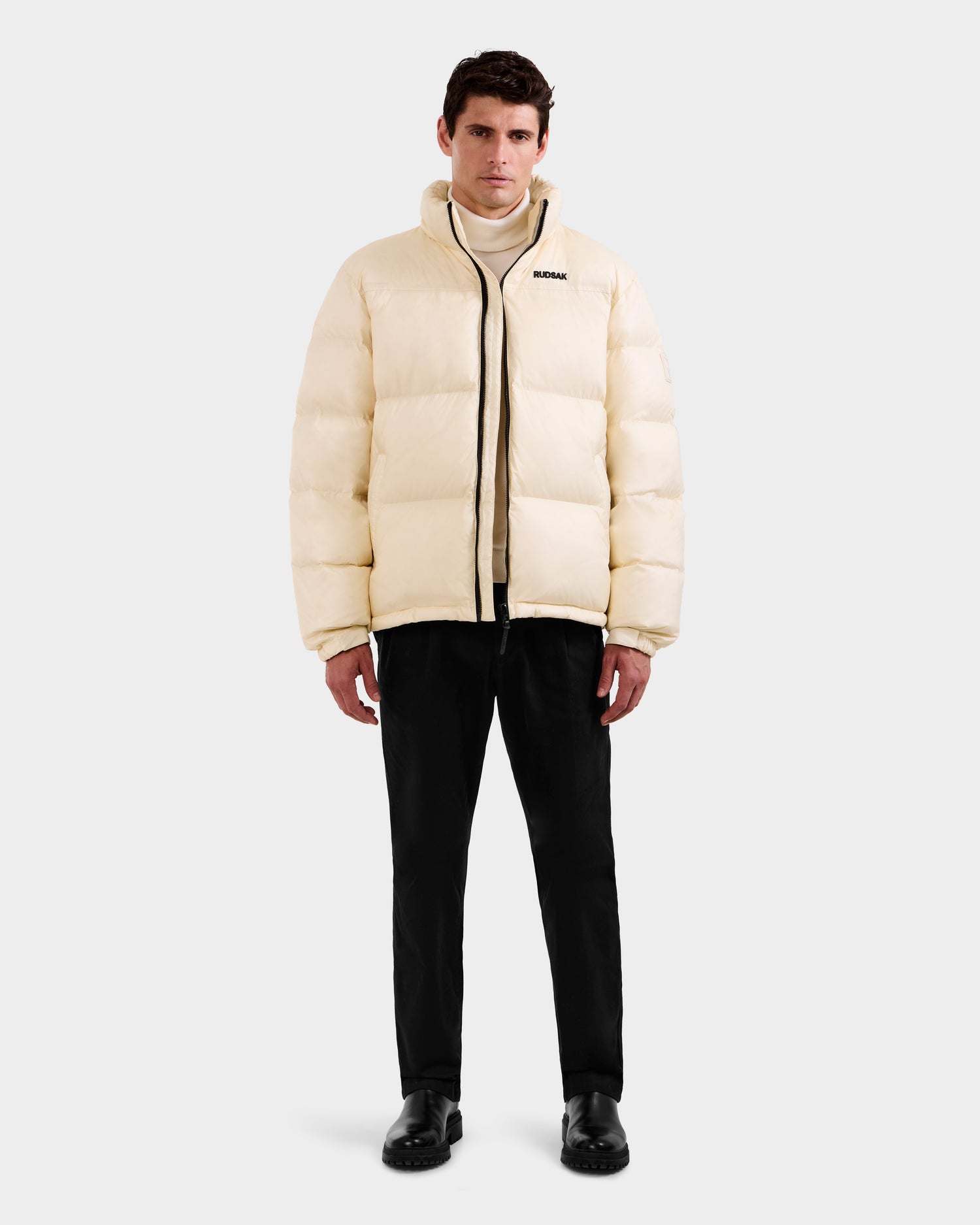 A man stands facing forward wearing the Rudsak STEFANO - 6125574 Vanilla men’s down puffer jacket, paired with black pants and shoes, against a plain light gray background.