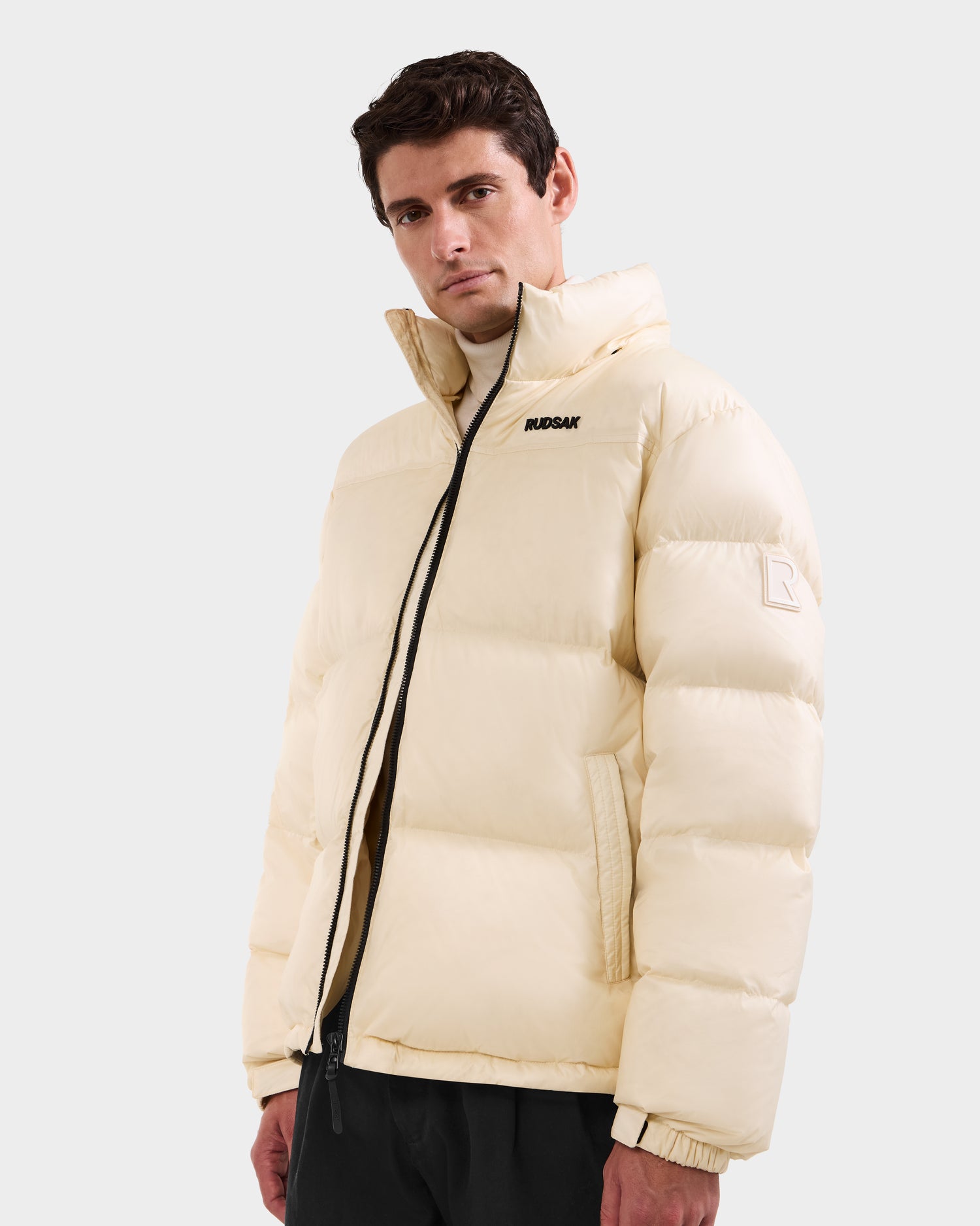 A man wears the Rudsak STEFANO - 6125574 Vanilla men’s down puffer jacket with a high collar and black zipper, featuring a small logo on the chest—ideal winter outerwear for chilly days against a light background.