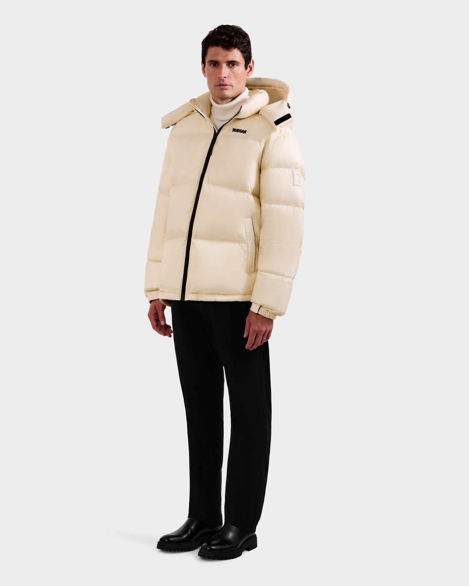 A man stands against a plain background wearing the Rudsak STEFANO - 6125574 Vanilla men’s down puffer jacket with a hood, chest logo, and front zipper, paired with black pants, black shoes, and a white turtleneck.