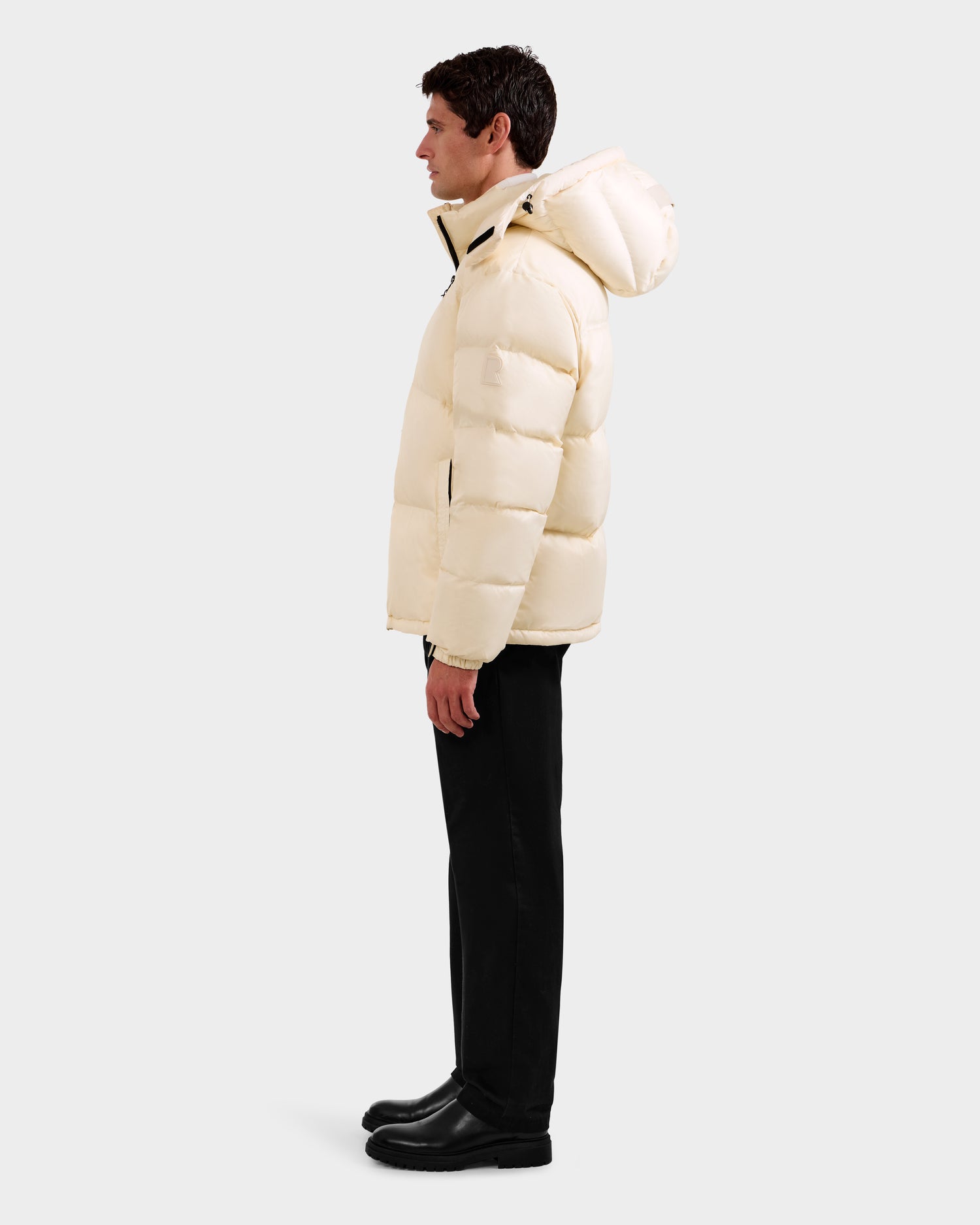 A man in profile wears the Rudsak STEFANO - 6125574 Vanilla men's down puffer jacket with a hood, paired with black pants and boots, set against a plain light gray background.