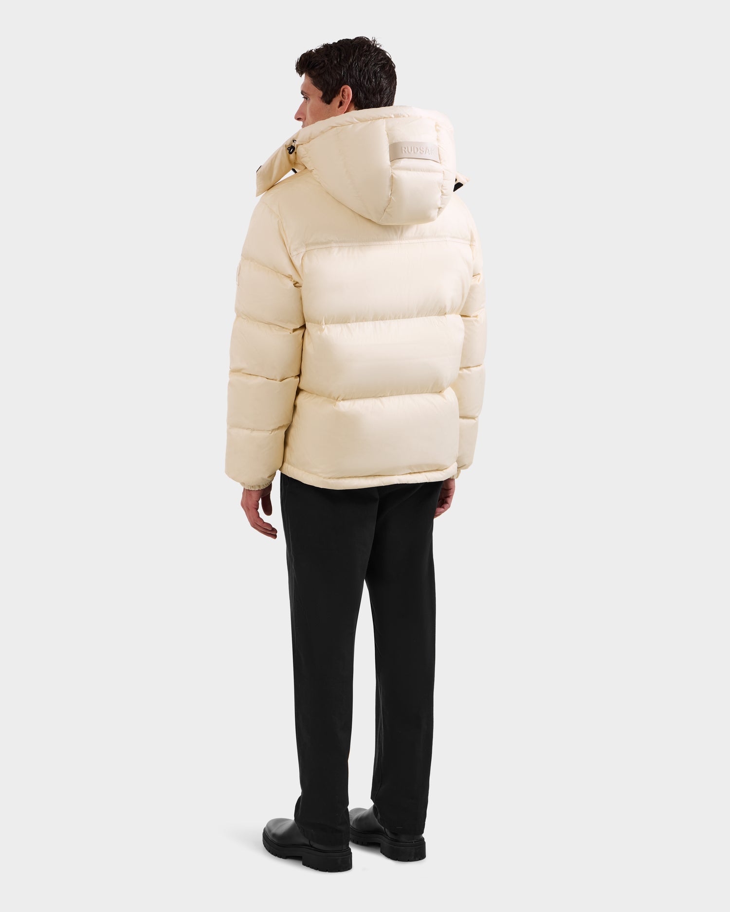 A person stands facing away, wearing a Rudsak STEFANO - 6125574 Vanilla men’s down puffer jacket with a hood, paired with black pants and shoes against a plain light background.