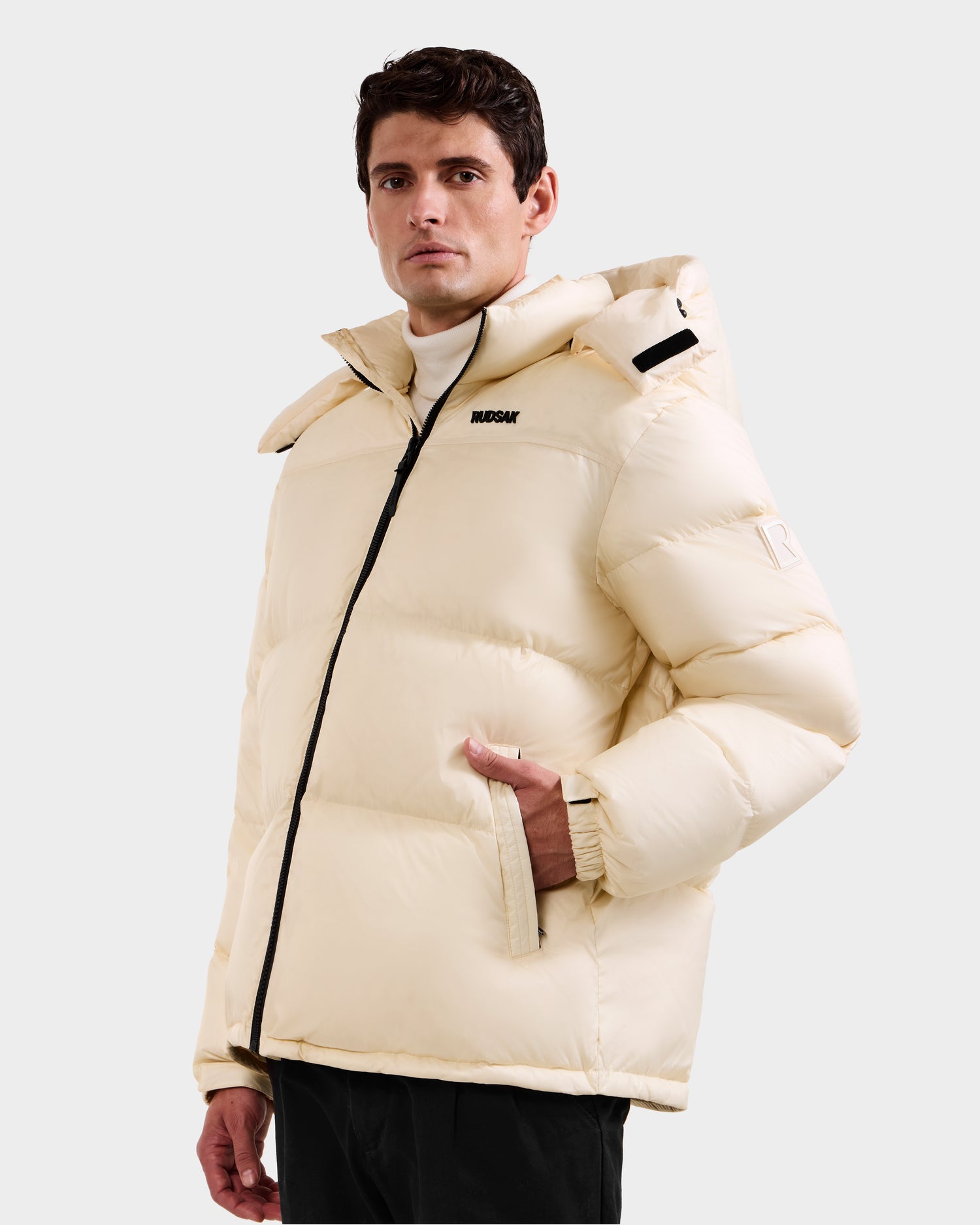 A man models the Rudsak STEFANO - 6125574 Vanilla, a cream quilted hooded jacket with a black zipper, standing against a light gray background—an ideal winter outerwear choice.