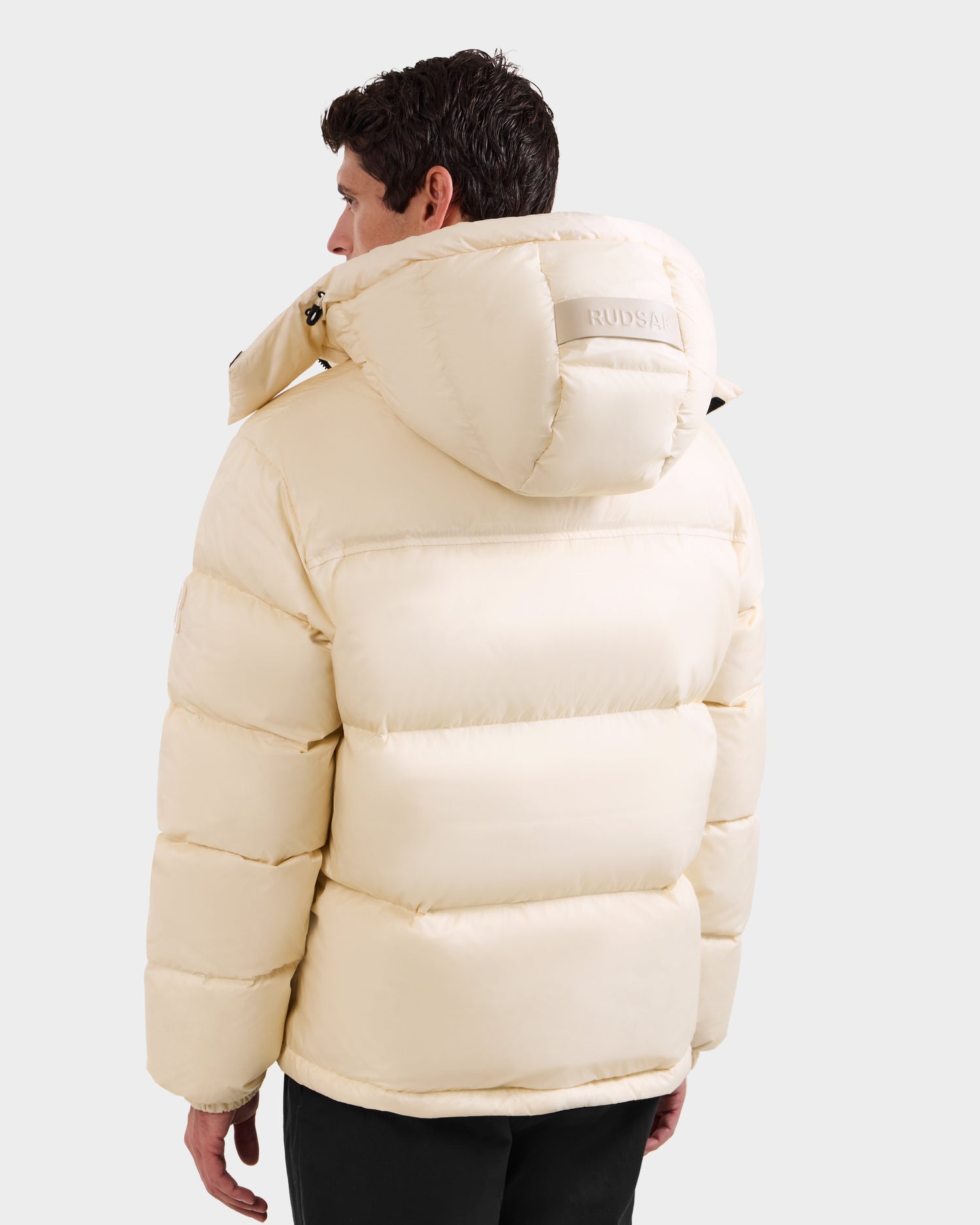 A man is seen from behind wearing the Rudsak STEFANO - 6125574 Vanilla men’s down puffer jacket with a hood. The light beige winter outerwear features horizontal stitching and a subtle brand logo on the hood, set against a plain background.