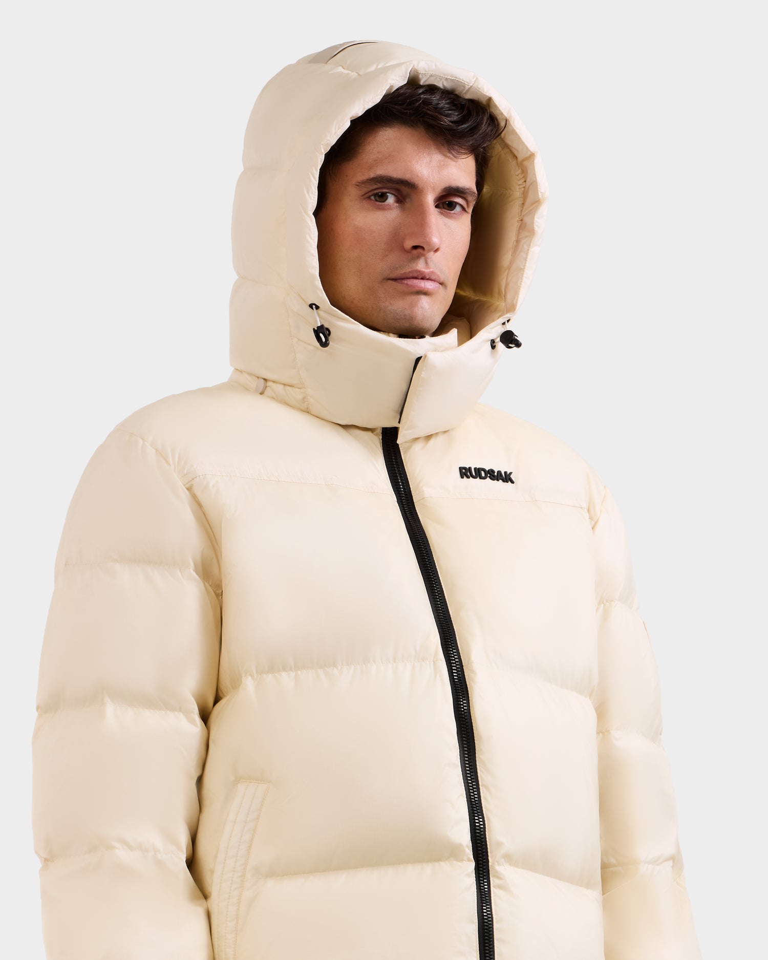 A man wears the STEFANO - 6125574 Vanilla men’s down puffer by Rudsak, featuring a hood and black zipper, with "RUDSAK" on the chest—posing against a light background in this stylish winter outerwear.