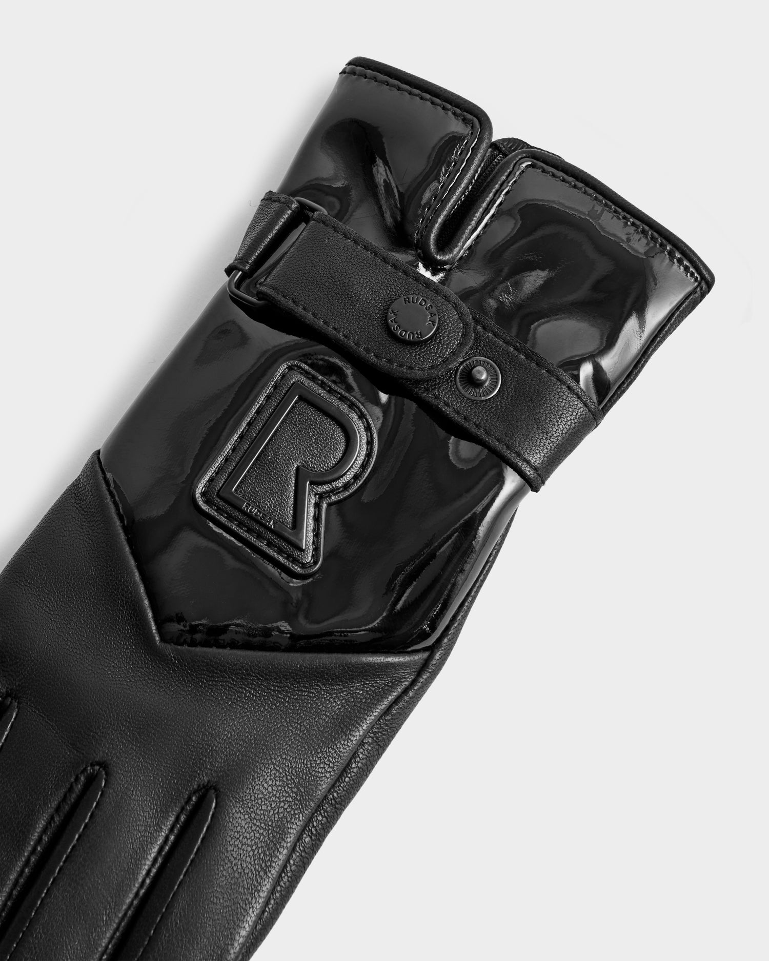 The Rudsak STROMBOLI - 8424509 Black women's leather glove features a shiny black finish, wrist strap with snap button, raised "R" on the back, and offers cashmere lining for added comfort, shown against a plain light background.