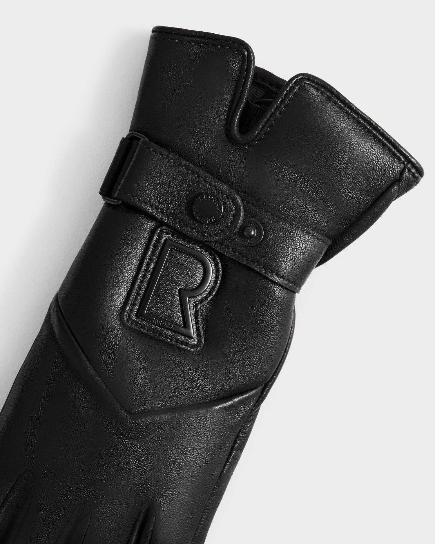 A close-up of Rudsak's STROMBOLI - 8424510 Black women's glove shows black leather, a Velcro strap, and an embossed R logo on the wrist. Touchscreen compatible, these gloves are both stylish and practical against a white background.