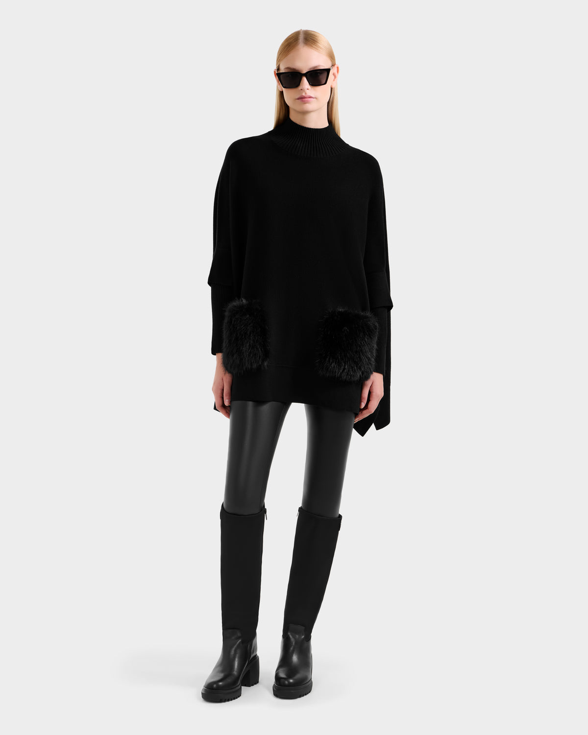 A woman in the Rudsak SWOON - 8525672 Black sweater poncho with faux fur pockets, black leather leggings, knee-high boots, and sunglasses stands against a plain light gray background.