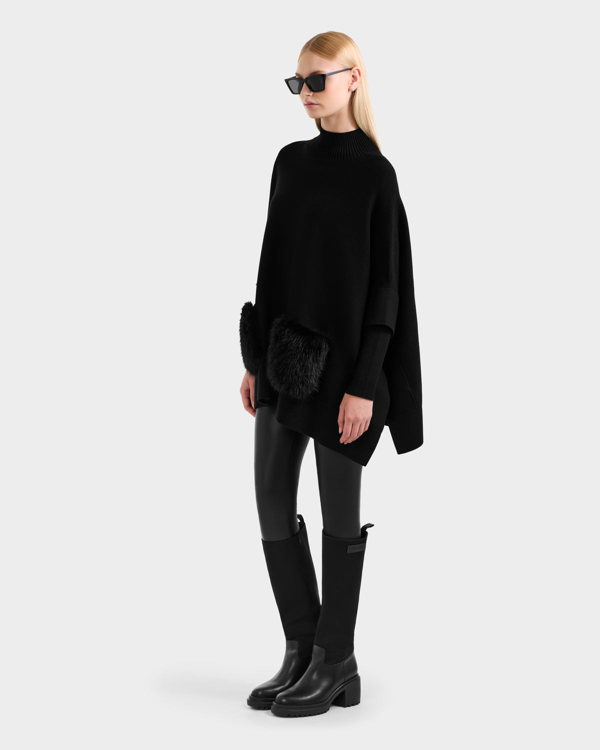 A woman in the Rudsak SWOON - 8525672 Black sweater poncho with faux fur pockets, styled with black leather pants and knee-high boots, embodies modern layering against a light gray background.
