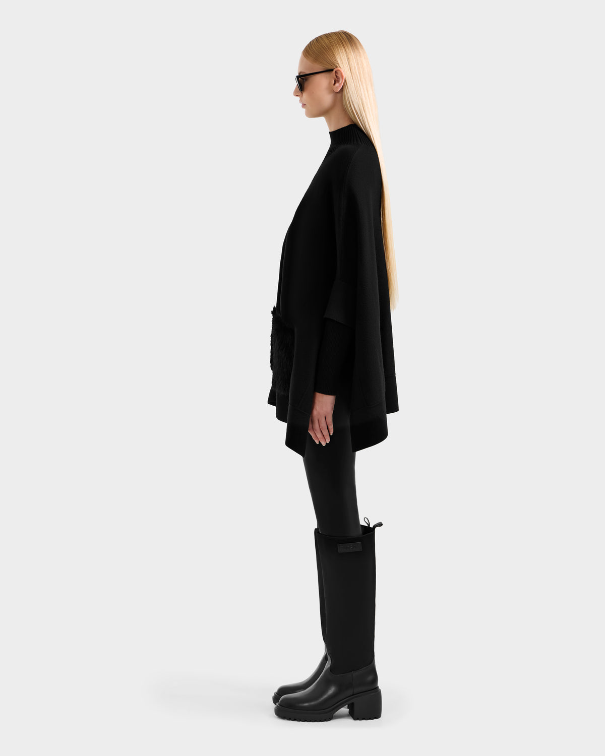 A woman with long straight blonde hair, sunglasses, and tall black boots stands in profile against a light gray background, wearing the Rudsak SWOON - 8525672 Black cape featuring a faux fur pocket and paired with black leggings.