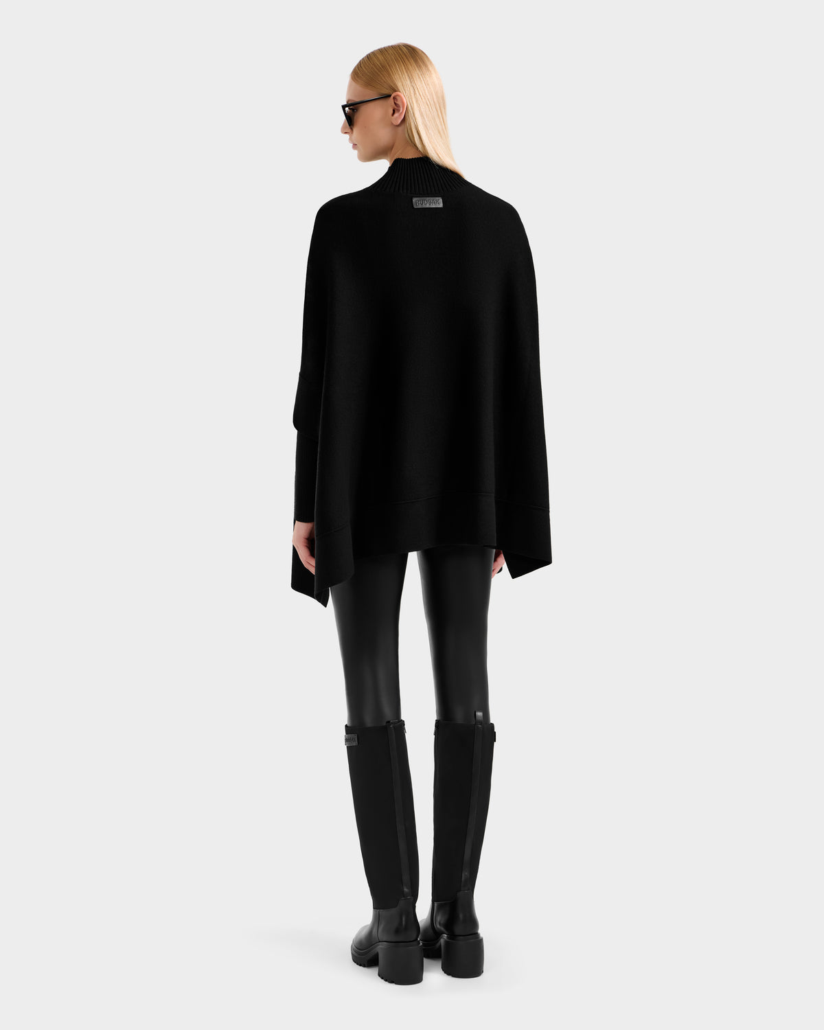 A woman with straight blonde hair and black sunglasses wears Rudsak’s SWOON - 8525672 Black faux leather leggings, a loose black sweater poncho, and knee-high boots, standing with her back to a light grey background.