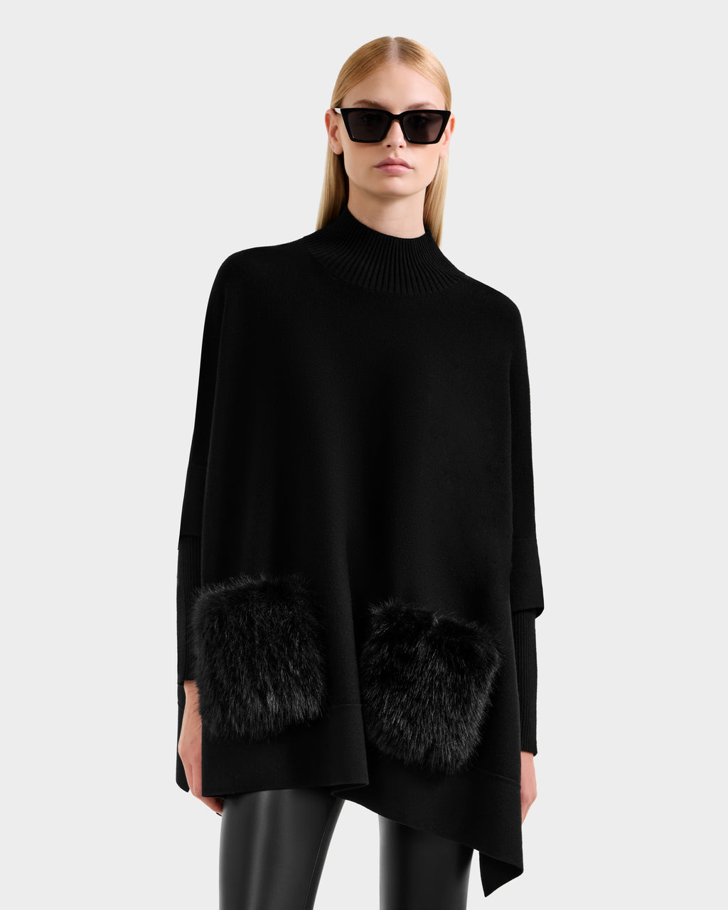 A woman with straight blonde hair models the Rudsak SWOON - 8525672 Black modern layering sweater poncho with two faux fur pockets, styled with black sunglasses and leather pants against a plain light background.