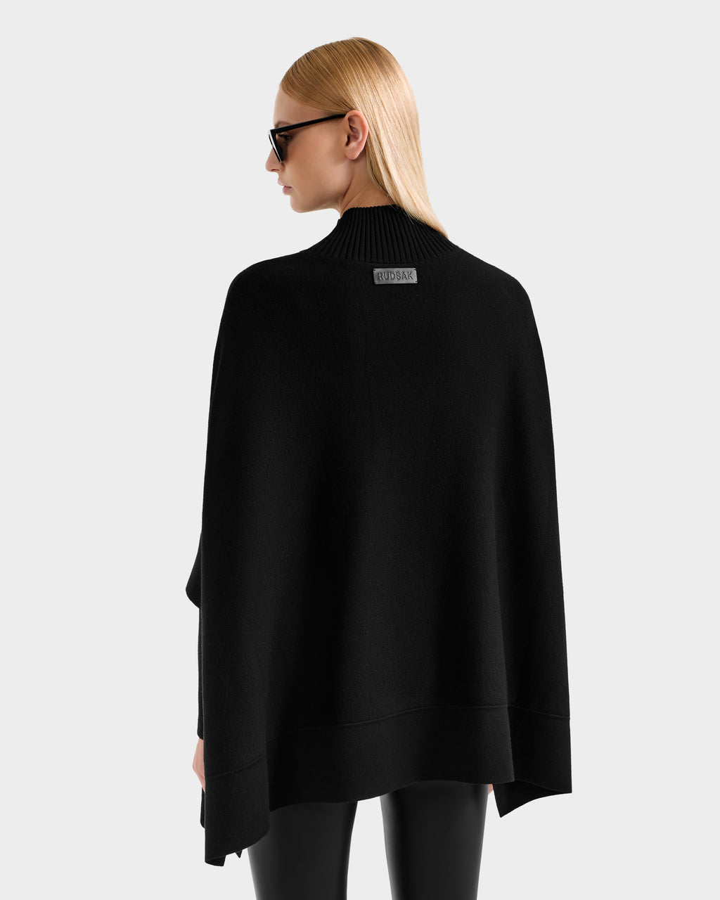 A person with straight blonde hair wears black sunglasses, black pants, and a Rudsak SWOON - 8525672 Black high-neck cape with modern layering. They are shown from the back against a plain light gray background.