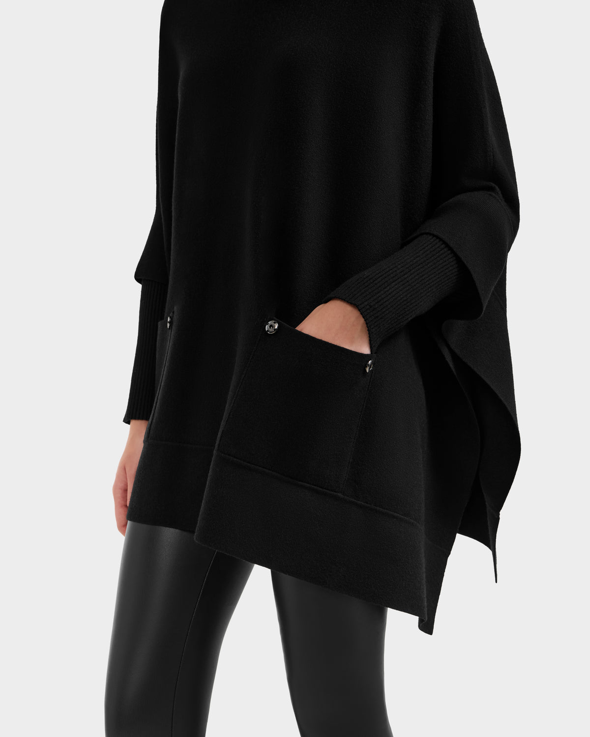 A person wearing Rudsak’s SWOON - 8525672 black sweater poncho with buttoned front pockets, long sleeves, and black leather-look leggings stands against a plain light background—ideal for modern layering.