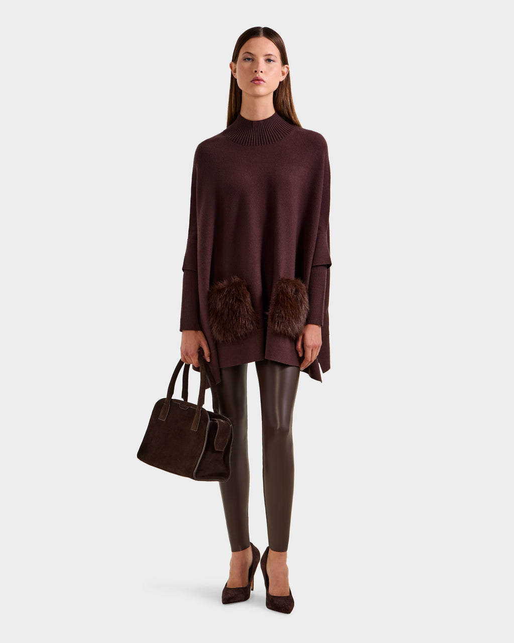 A woman poses against a plain background wearing the Rudsak SWOON - 8525672 Mocha sweater poncho with faux fur pockets, paired with shiny brown leggings, brown heels, and a matching brown handbag at her side.