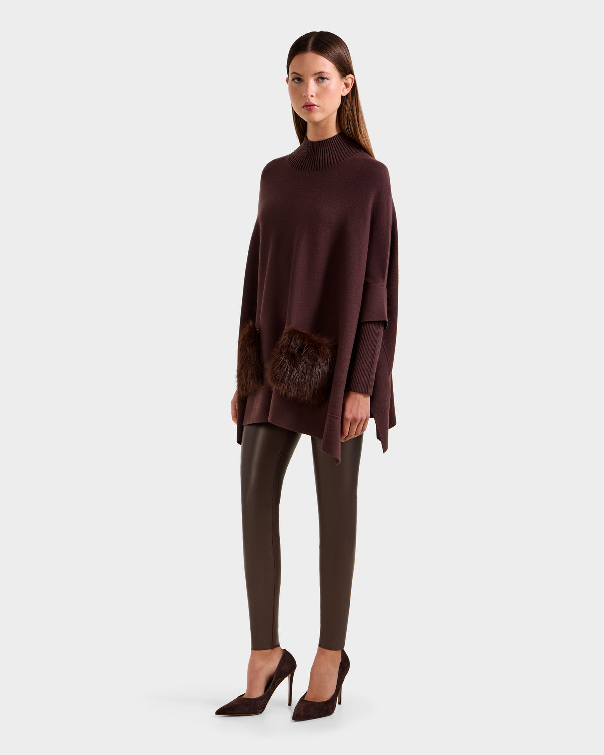A woman wears the Rudsak SWOON - 8525672 Mocha, a loose dark brown sweater poncho with faux fur pockets, paired with fitted brown pants and dark heels—an elegant modern layered look set against a plain light background.