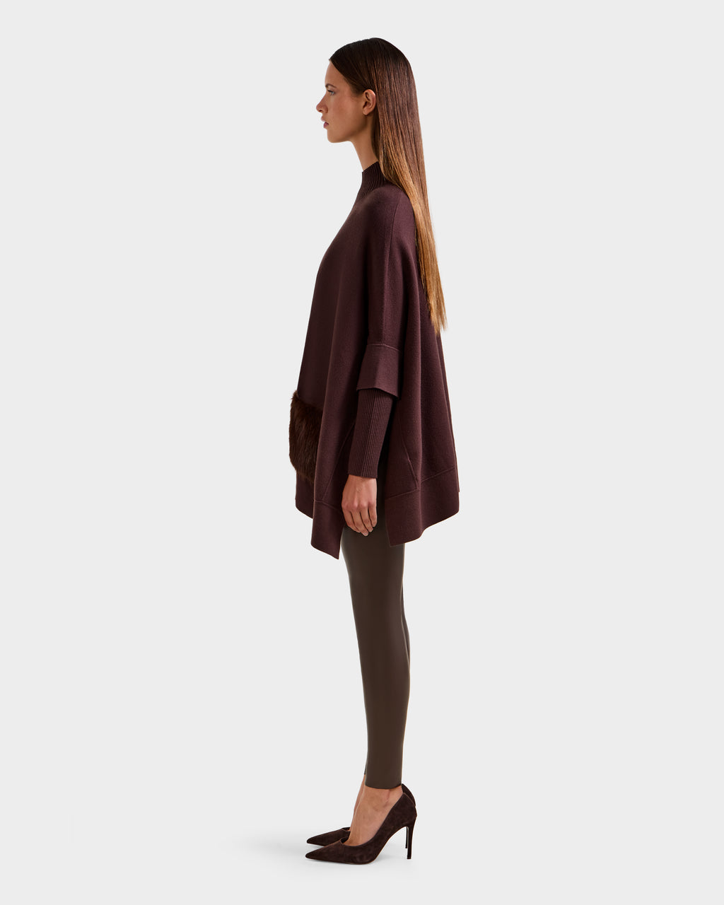 A woman in profile poses against a plain background, wearing the Rudsak SWOON - 8525672 Mocha sweater poncho with modern layers, dark leggings, and brown heels. Her straight hair is down and her arms are relaxed at her sides.