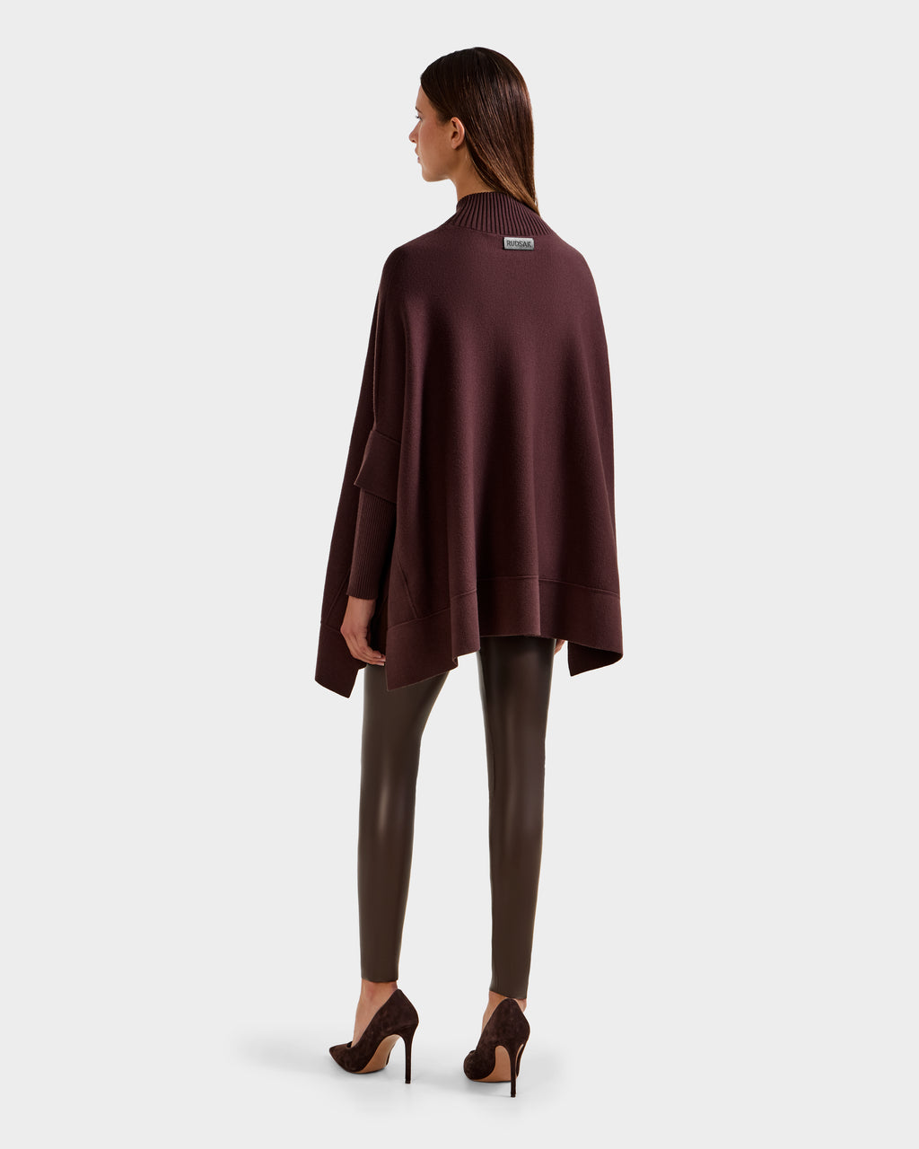 A woman with straight hair stands facing away, wearing the Rudsak SWOON - 8525672 Mocha sweater poncho over matching fitted pants and high-heeled shoes, set against a plain light gray background.