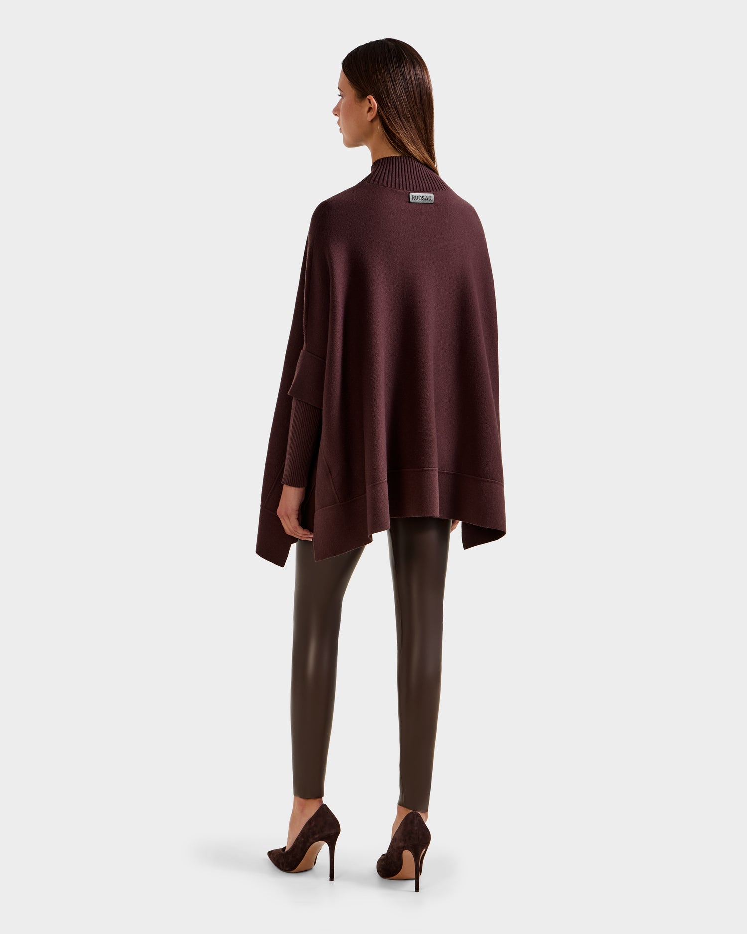 A woman with straight hair stands facing away, wearing the Rudsak SWOON - 8525672 Mocha sweater poncho over matching fitted pants and high-heeled shoes, set against a plain light gray background.