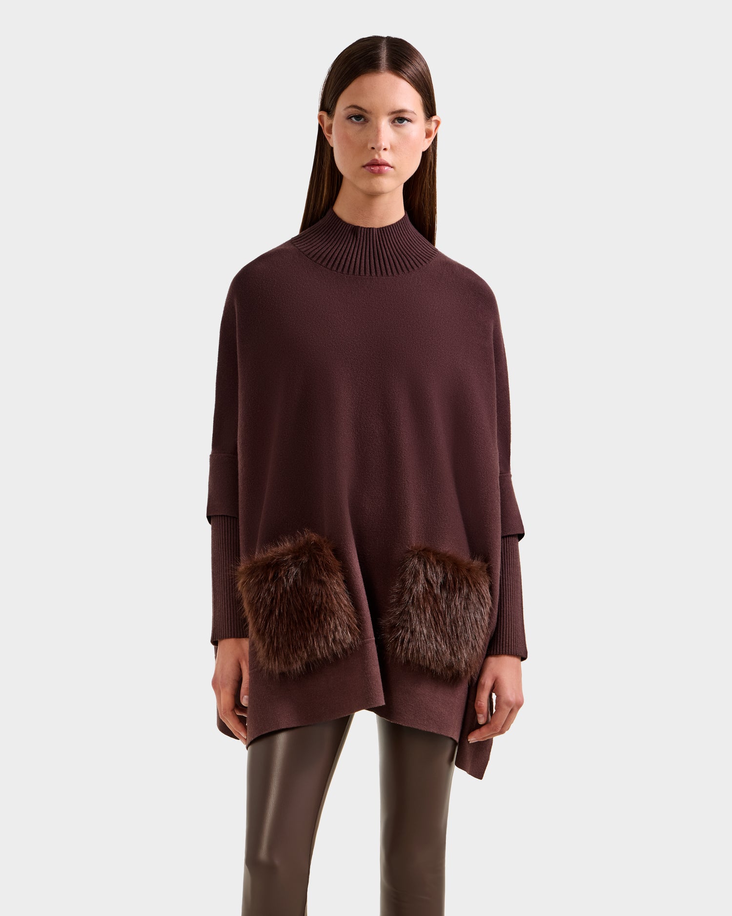 A woman models the Rudsak SWOON - 8525672 Mocha: a loose, brown knit sweater with a high ribbed collar, long sleeves, and two faux fur pockets—ideal for layering—paired with shiny brown leggings against a simple light backdrop.
