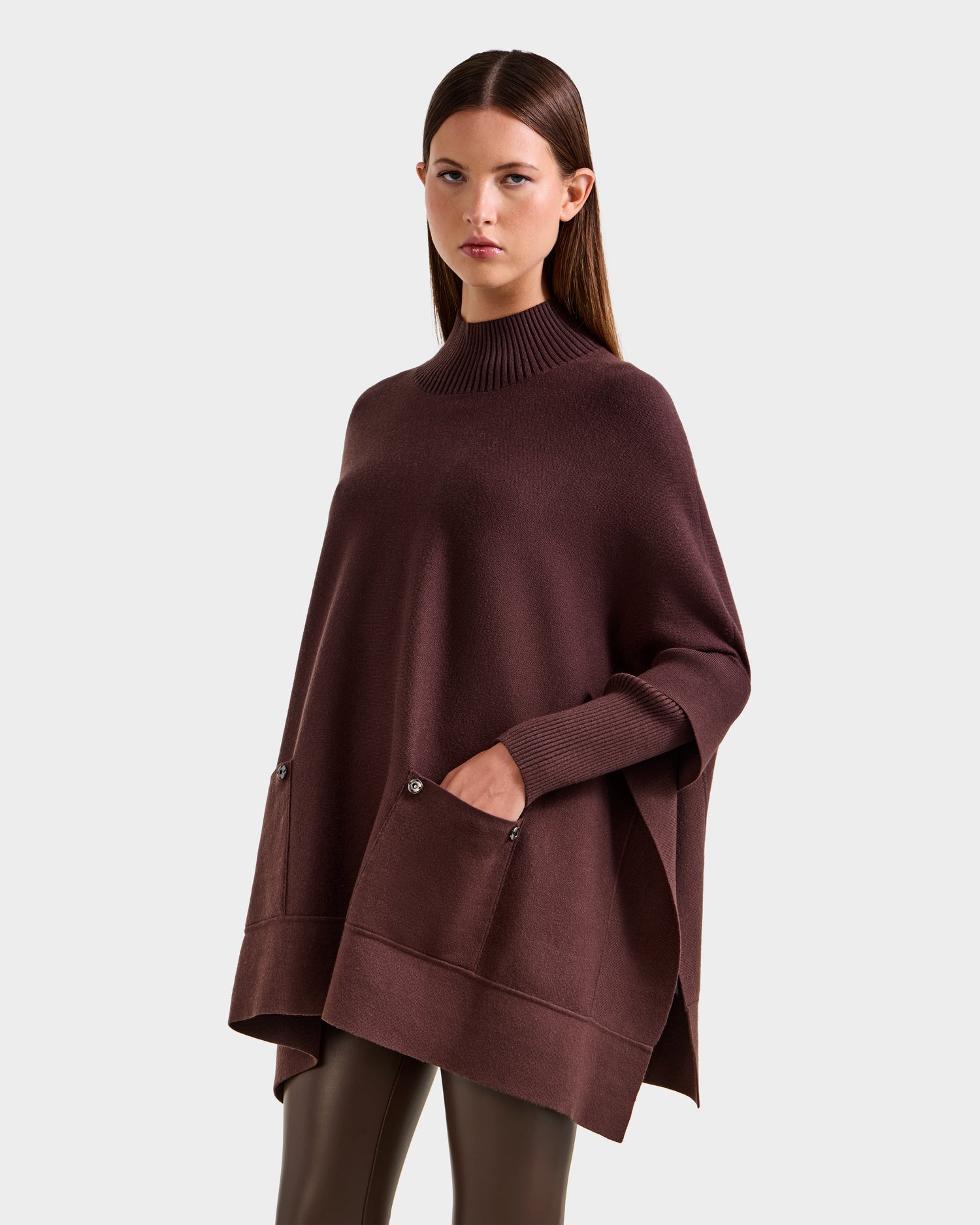 A woman with straight brown hair models the Rudsak SWOON - 8525672 Mocha poncho, featuring a loose fit, ribbed turtleneck, and front pockets, paired with shiny brown leggings against a plain light gray background.