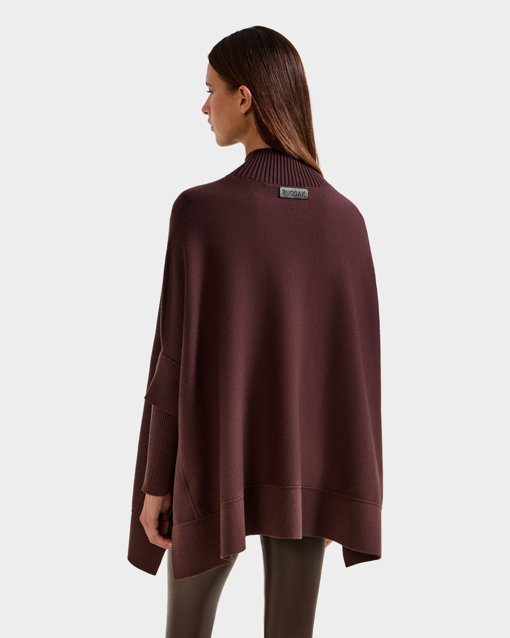 A woman with straight brown hair models the Rudsak SWOON - 8525672 Mocha sweater poncho, featuring a high ribbed collar and loose fit, standing with her back to the camera against a plain light gray background—ideal for modern layering.