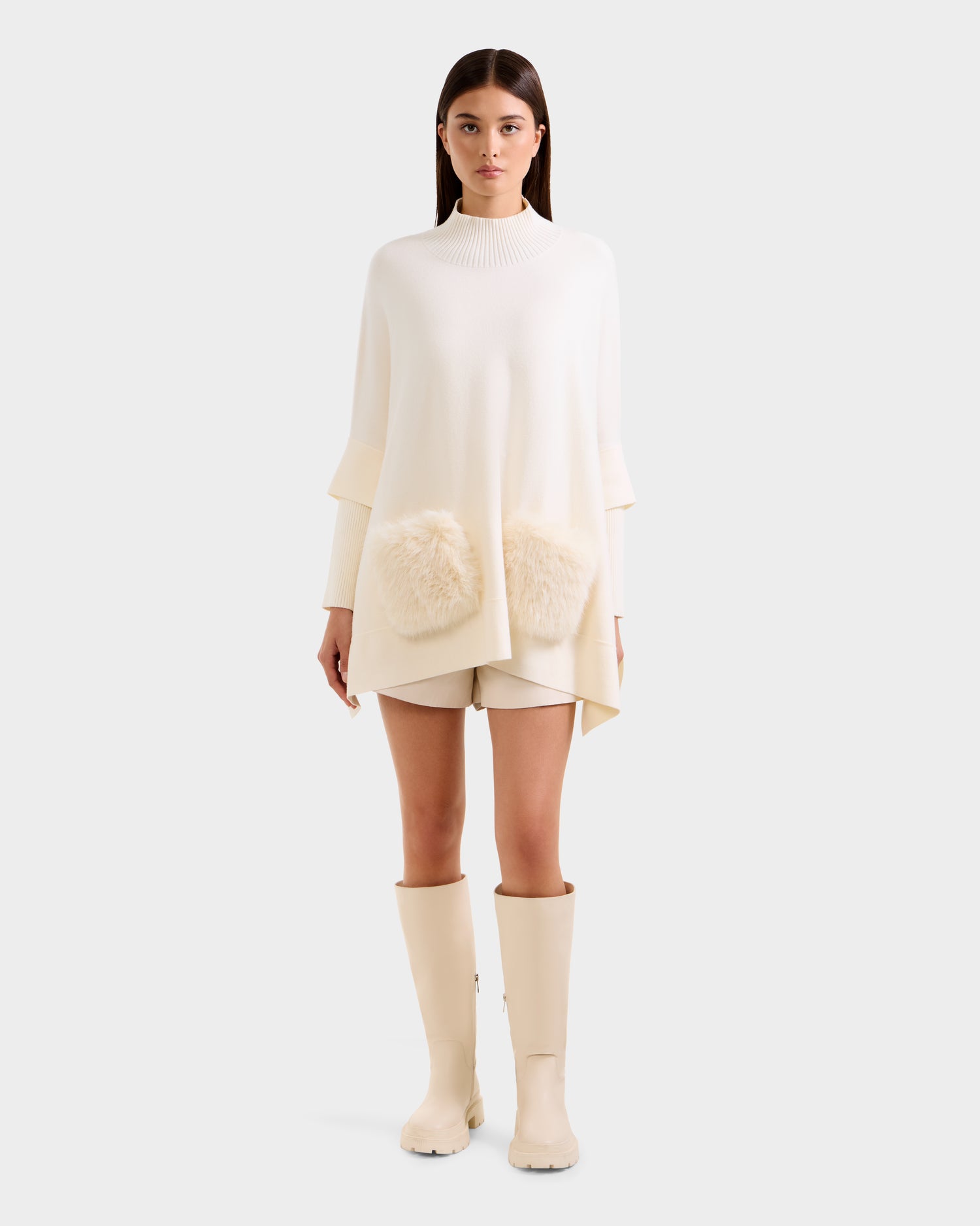 A woman faces forward in the Rudsak SWOON - 8525672 Vanilla sweater poncho with faux fur pockets, matching shorts, and cream knee-high boots. She has straight brown hair and a neutral expression against a plain white background.