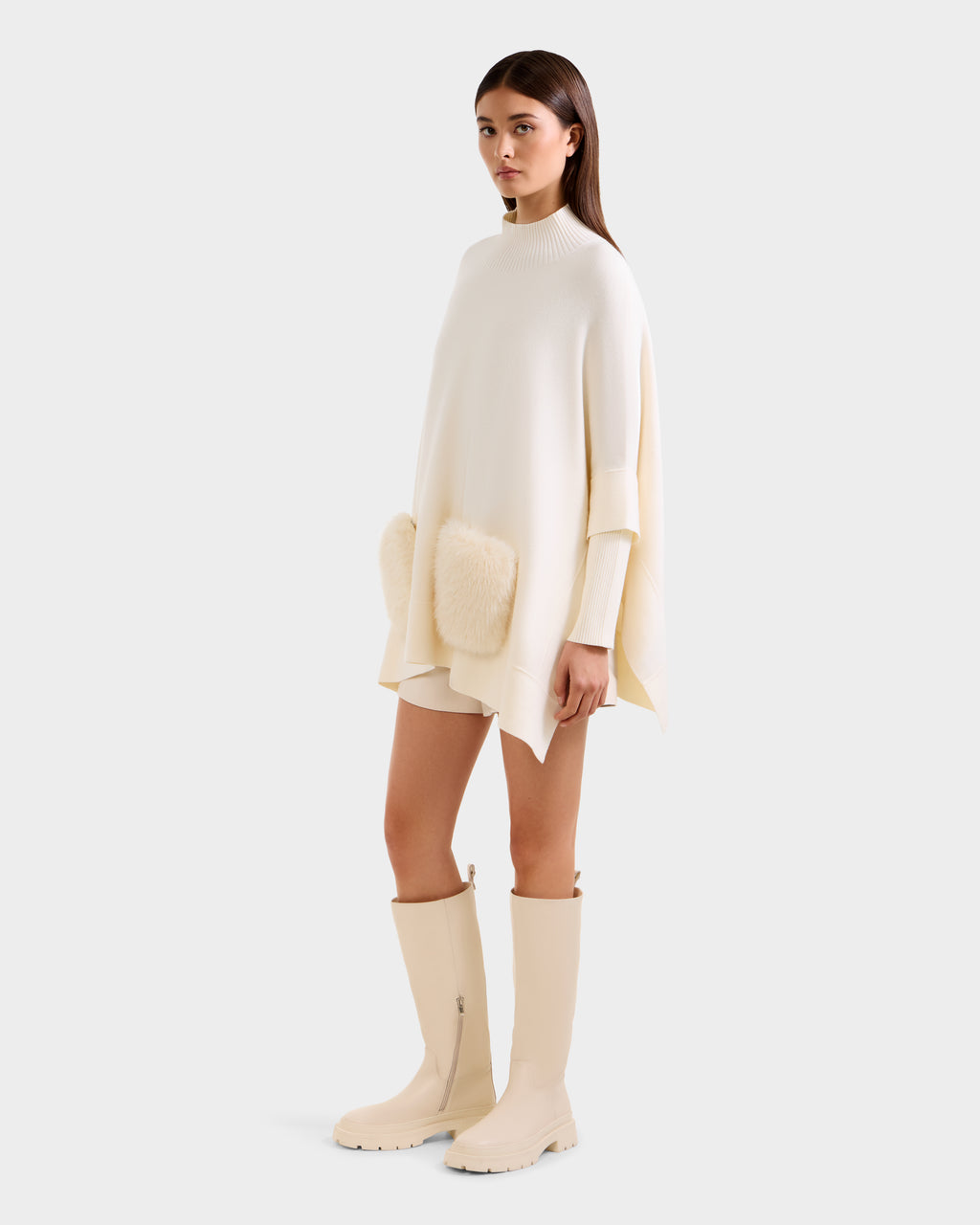 A woman models the Rudsak SWOON - 8525672 Vanilla, an oversized cream sweater poncho with faux fur pockets, styled with matching shorts and knee-high boots for a chic, modern layered look against a light background.