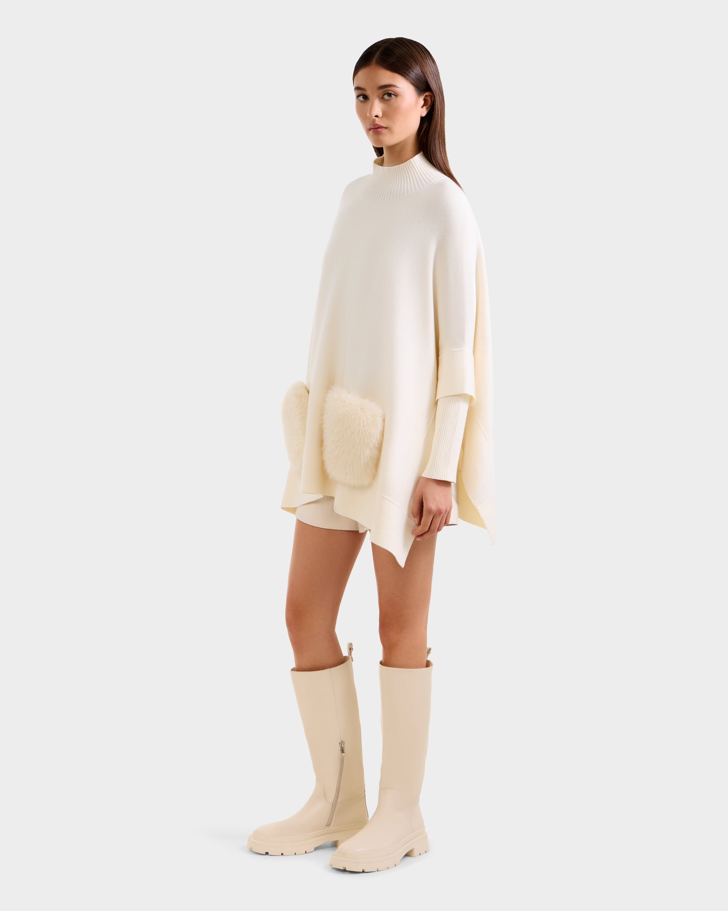 A woman models the Rudsak SWOON - 8525672 Vanilla, an oversized cream sweater poncho with faux fur pockets, styled with matching shorts and knee-high boots for a chic, modern layered look against a light background.